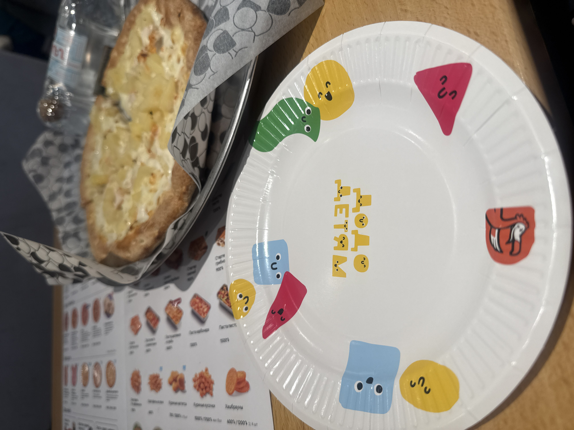 The image presents a paper plate with cartoonish shapes printed around its edge, situated on a wooden table. Adjacent to the plate is a metallic container filled with what appears to be bread covered in a light-colored spread. The bread is partially obscured by patterned paper. In the background, a menu board displays various food items and prices. The Cyrillic script on the menu, combined with the context of "Dodo Детям" on the plate, suggest this image was taken at a Dodo Pizza location, a chain with a presence in Yerevan, Armenia.