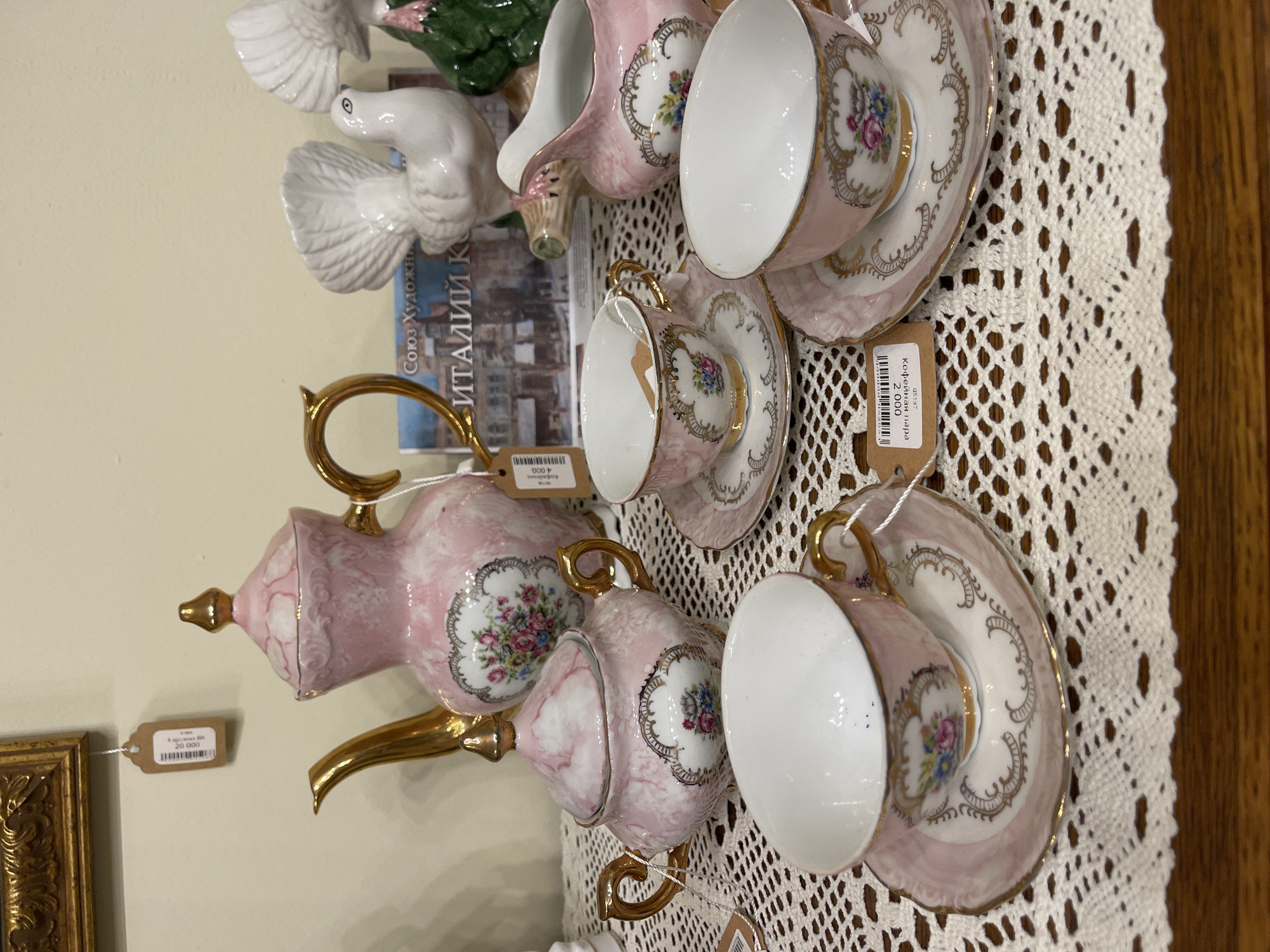 The image displays a collection of vintage pink and gold porcelain teacups and saucers, a teapot, a sugar bowl, and a creamer, all adorned with floral patterns. These items are arranged on a white lace doily atop a wooden surface. In the background, several white ceramic bird figurines are visible, along with a book featuring a European cityscape. A framed picture hangs on the wall above the display. Price tags are attached to some of the items, with one tag clearly legible indicating "Кофейная пара" (Coffee pair) and a price of "2 000". The overall aesthetic suggests an antique shop or a curated display.
The tag indicating "Кофейная пара" and a price of 2000, along with the likely context of an antique or collectible item, suggests this photograph was taken in an antique shop in the city of Svetlogorsk, Russia. Such items are commonly sold in antique shops, and Svetlogorsk, like many Russian cities, has such establishments.