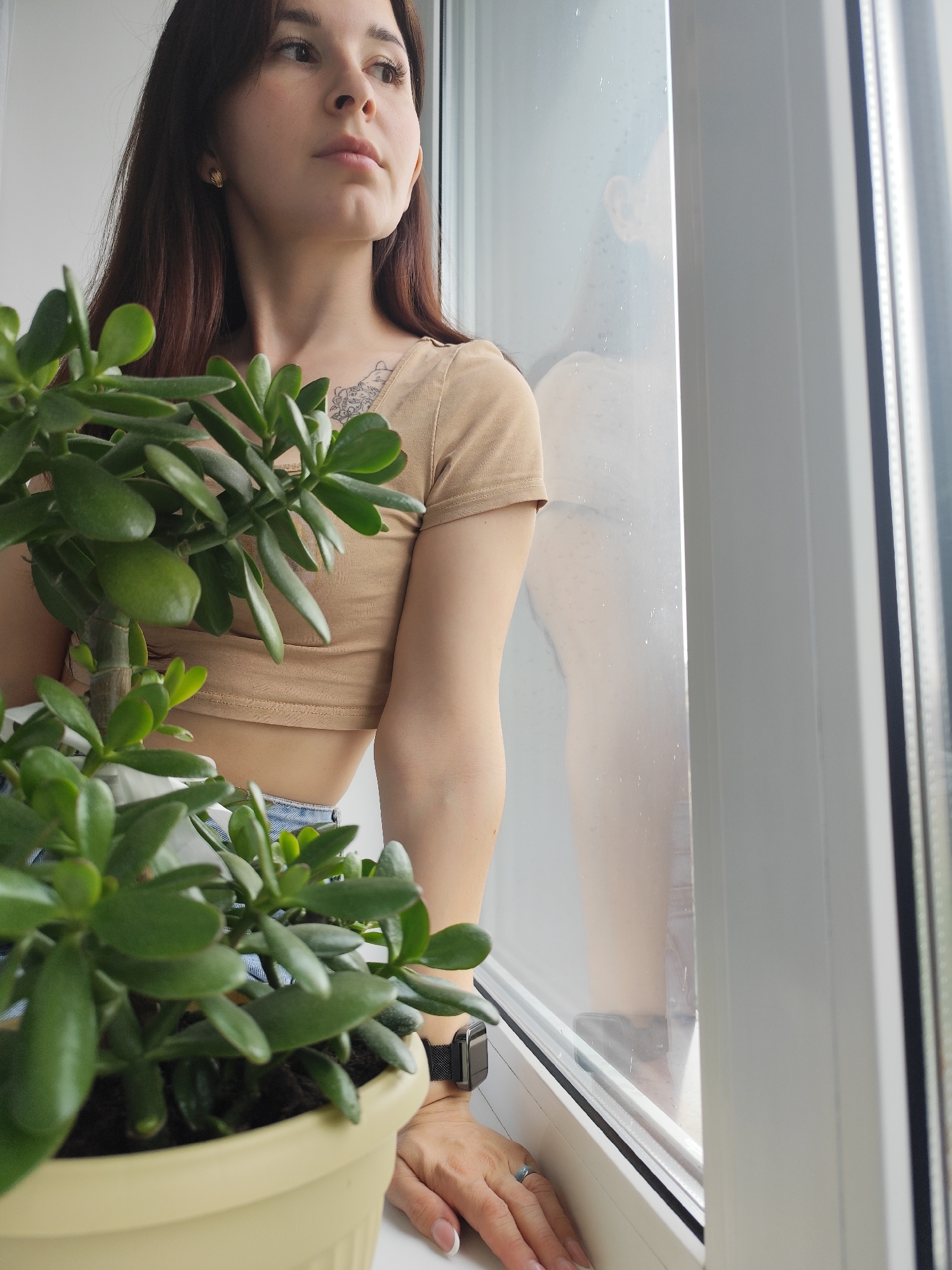 A young woman with long, dark hair stands by a window, gazing thoughtfully into the distance outside. She is positioned on the left side of the frame, with her profile visible. Her left hand rests gently on the white windowsill, adorned with a silver-toned ring on her ring finger and a dark smartwatch on her wrist. She wears a beige, short-sleeved cropped top, which reveals a tattoo on her upper right chest/shoulder area, and what appear to be denim shorts or jeans. A small golden earring is visible in her right ear.
In the foreground, a large potted jade plant (Crassula ovata), also known as a money tree, occupies the lower left corner and extends upwards, partially obscuring the woman's lower body and right arm. The plant is in a light yellow pot, with vibrant green, fleshy leaves.
The windowpane shows numerous water droplets, suggesting it is raining or has recently rained outside. A blurred reflection, appearing to be the woman's own, is visible on the window's surface to her right. The window frame is white and modern. The overall scene conveys a calm, reflective moment indoors, with soft, natural light illuminating the space. There is no visible text in the image. The setting is an indoor space, located in Фоки, Russia.