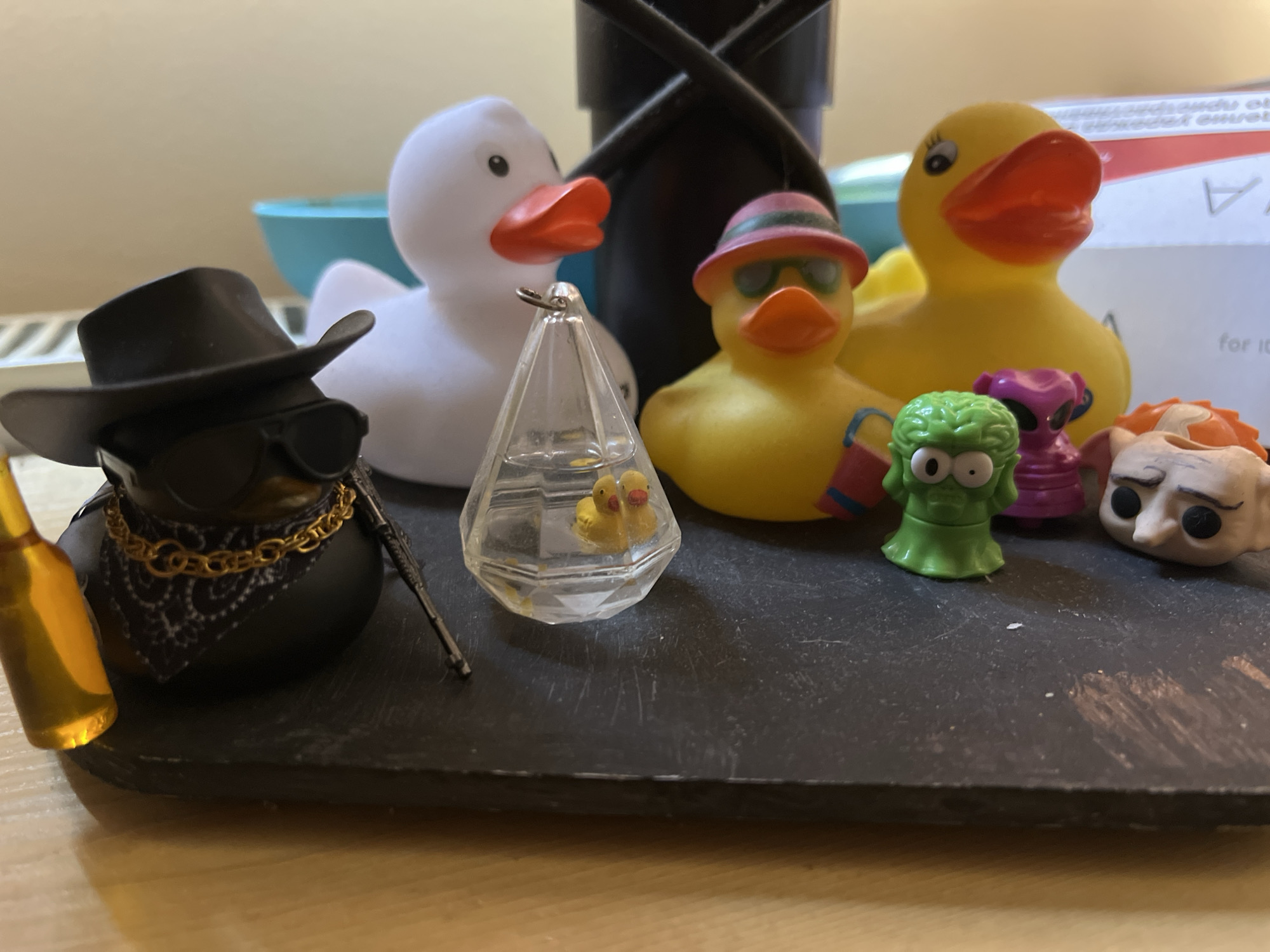 A still life arrangement of several rubber ducks and figurines is depicted on a dark, textured surface. From left to right, a black rubber duck wearing a cowboy hat, sunglasses, a gold chain, and a bandana is positioned next to a small golden bottle. To its right, a clear, faceted container holds two tiny yellow rubber ducklings. Behind these, a large white rubber duck stands. Further to the right, a yellow rubber duck wearing a pink and green striped hat and sunglasses is present. Next to it, another large yellow rubber duck is visible. In the foreground, near the right side of the image, are three small figurines: a green, brain-like creature, a purple alien-like figure, and a pale, grotesque face with dark eyes and hair. The background includes a blurred blue bowl and part of a cardboard box with text that is partially legible. The lighting suggests an indoor setting, possibly on a table. There is no indication of Sofia, Bulgaria, in the image.