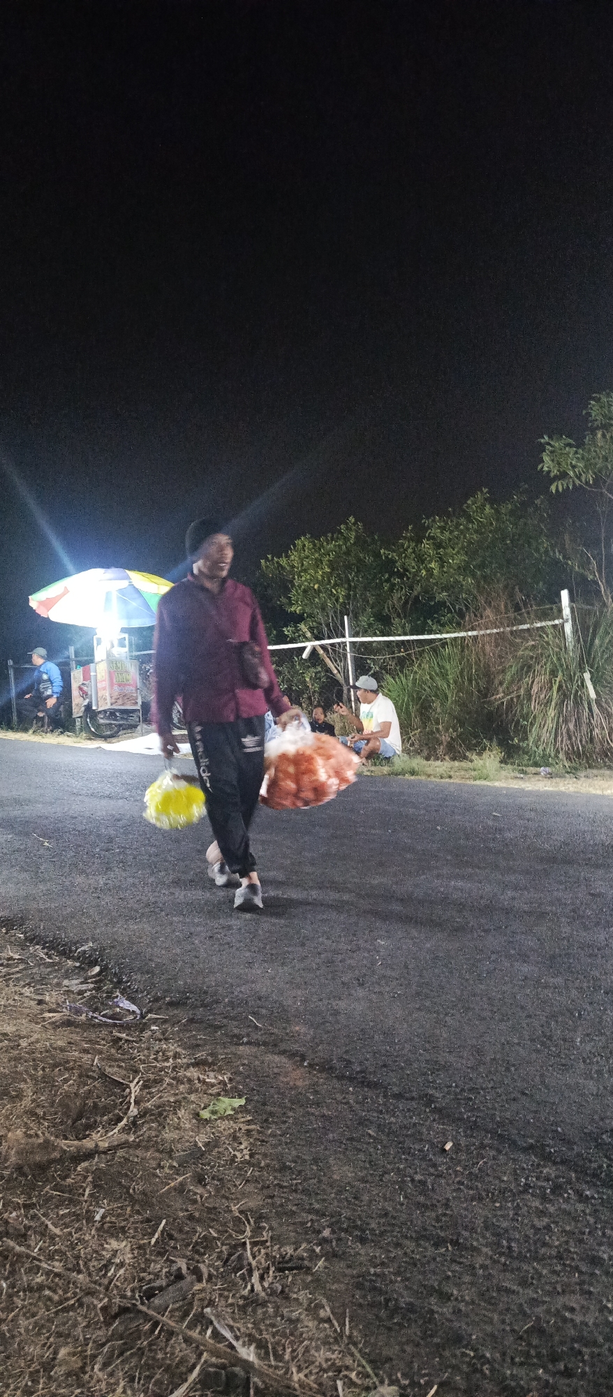 A person walks down a paved road at night in Sumbersekar, Indonesia, carrying two large, round bags of what appear to be food items. The person is wearing a dark beanie, a maroon collared shirt, and dark pants with white stripes. They are also wearing dark shoes. In their right hand, they carry a yellow, fluffy object, possibly a toy or decoration, and in their left hand, they carry a large, reddish-orange bag.
In the background, there is a food cart with a colorful umbrella illuminated by lights. To the left of the cart, a person in a blue jacket is seated. Further to the right, a group of people are sitting on the ground near some vegetation and a white fence. One individual, wearing a light-colored t-shirt and shorts, is looking towards the person walking. The environment suggests a roadside setting with trees and tall grass. The ground on the left side of the road appears to be unpaved and covered with debris. The sky is dark, indicating nighttime.