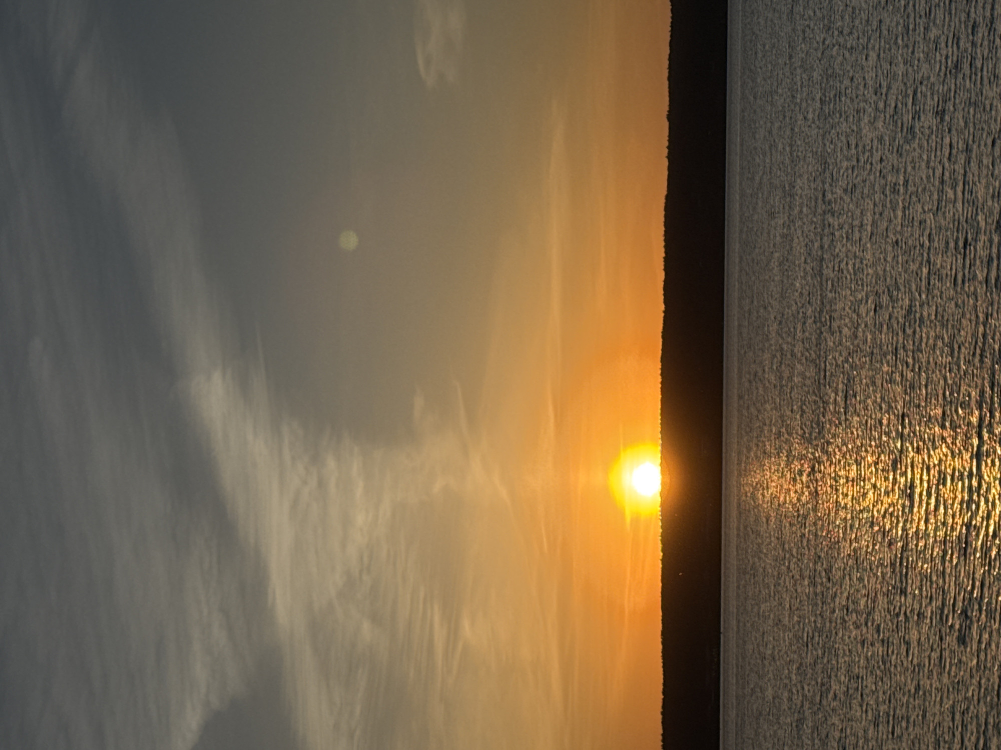 A sunset over water is depicted. The sun is on the horizon, with a small strip of land cutting between the sky and the water. The sun’s light reflects off of the water. The sky is a pale yellow where the sun is. A gray-white cloud is in the sky. The scene appears to be in the city of Unknown, Unknown.