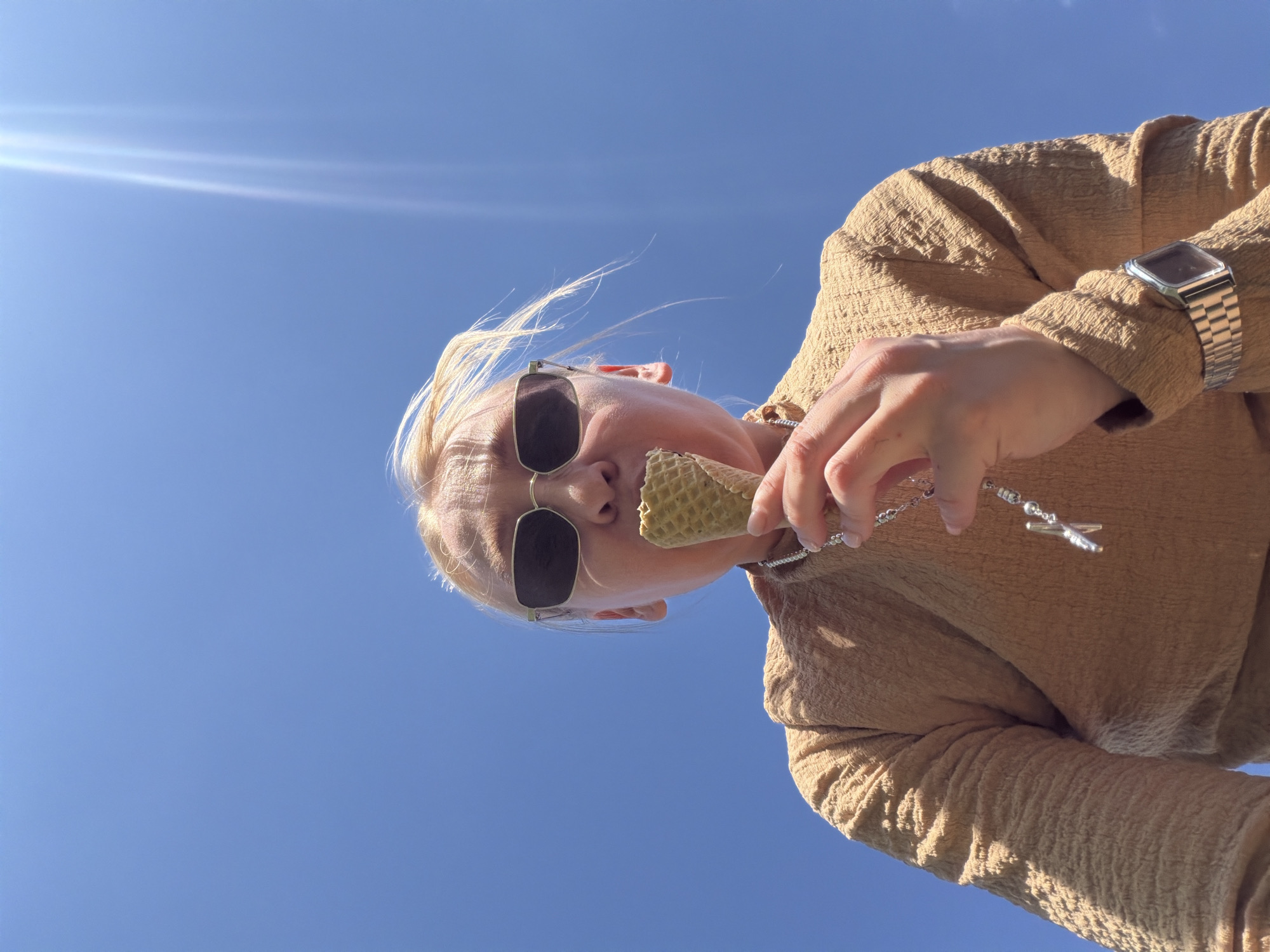 A low-angle shot depicts a person outdoors against a clear blue sky with a visible sun glare. The person has fair skin, blond hair, and is wearing sunglasses. They are holding an ice cream cone in their right hand. The person is wearing a textured tan or brown shirt and a silver wristwatch. A silver necklace with a pendant is visible. The location is unspecified, and there is no information connecting it to the city of Chaikovsky, Russia.