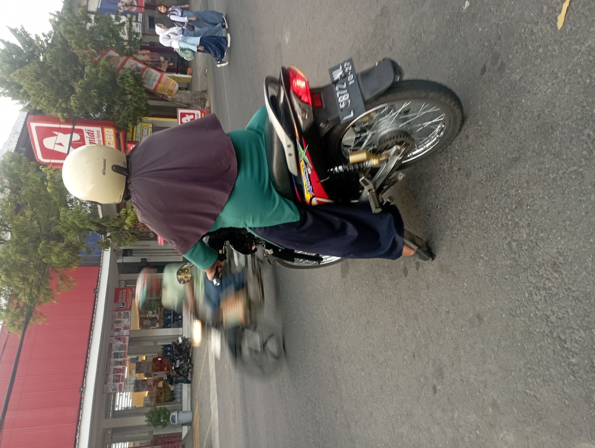 The image shows a street scene in Ganung Kidul, Indonesia. In the foreground, a person wearing a green hijab and a light yellow helmet is riding a motorcycle. The motorcycle has a license plate that reads "L 5872 NL" and a sticker with "10-27". The rider's legs are covered by a long, dark blue garment. In the background, a red building with white trim and storefront windows is visible. A sign above the storefront reads "A Nidi". Further down the street, two young women in school uniforms, also wearing hijabs, are walking. Another person on a motorcycle is blurred in motion to the left of the frame. The street is paved with asphalt, and trees line the area.