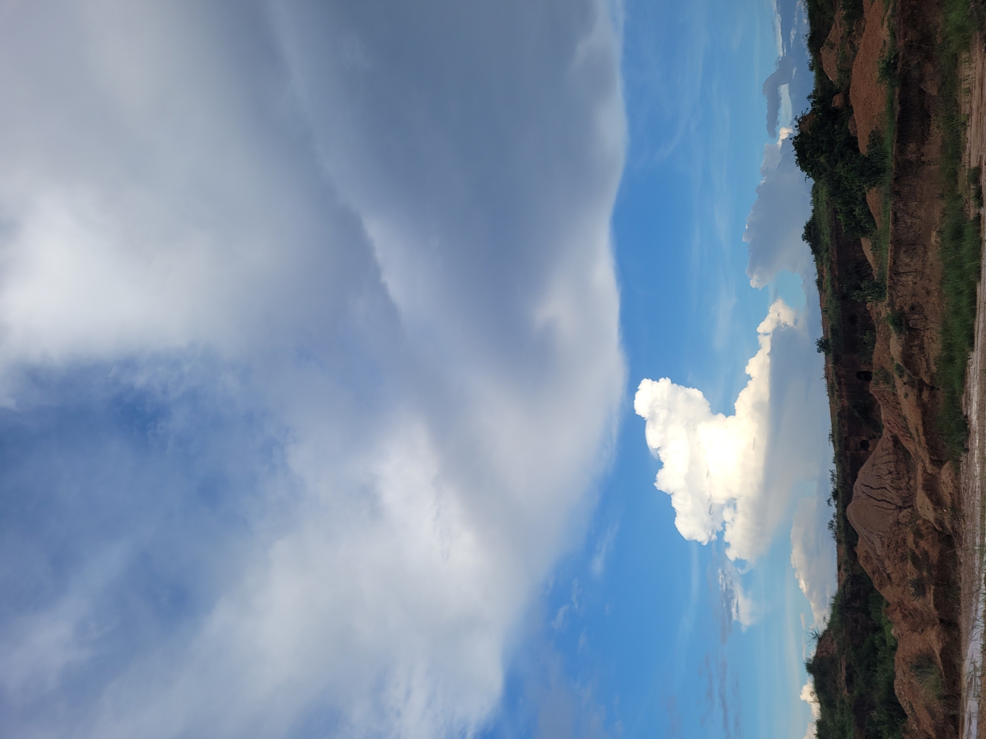 The image depicts a landscape with a focus on the sky and a rough, eroded terrain. In the upper portion of the image, a dramatic sky features a mixture of white and gray clouds against a bright blue sky. The clouds are varied in texture and form, with some appearing wispy and scattered, while others are dense and stormy.
The lower portion of the image shows a barren, hilly landscape characterized by exposed soil and sand. The ground is uneven, with steep banks and mounds of reddish-brown earth. Sparse green vegetation, including grasses and a few scattered bushes and trees, grows on the slopes and along the crest of the hills. In the foreground, there is a hint of water, possibly a shallow pool or muddy ground. The overall impression is one of a dry, possibly arid or semi-arid environment that has undergone significant erosion.
The scene appears to be an outdoor location, likely a natural or semi-natural area. Without further information or context, it is impossible to definitively place this image in Bukuru, Nigeria, as it represents a generic landscape that could be found in various regions with similar geological and climatic conditions. However, if this is indeed Bukuru, it suggests an area with exposed geological formations and a predominantly clear or partly cloudy sky.