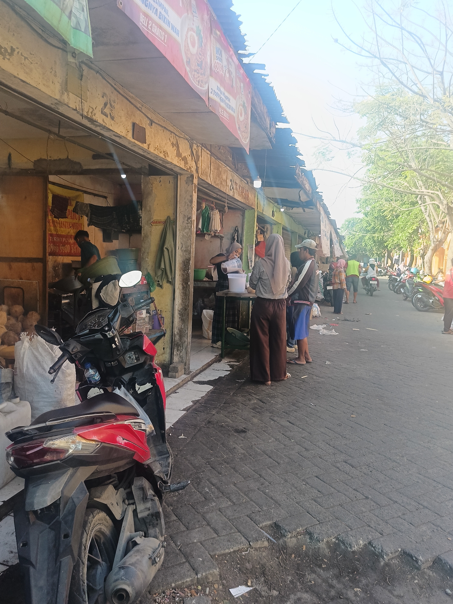 The image depicts a street scene in Ketajen, Indonesia, likely a market area. In the foreground, a red and black scooter is parked on a paved street. The street is lined with open-air shops and stalls. One stall on the left features a man in a green shirt attending to large green buckets and what appear to be coconuts. A banner with text in Indonesian is visible above this stall. Further down the street, several people are gathered, some standing and some walking. There are more stalls and shops visible, with awnings and corrugated metal roofs. Various items are displayed, including clothing and what might be food items. In the background, trees line the street, and several motorcycles are parked. The overall impression is of a busy, working-class commercial district.