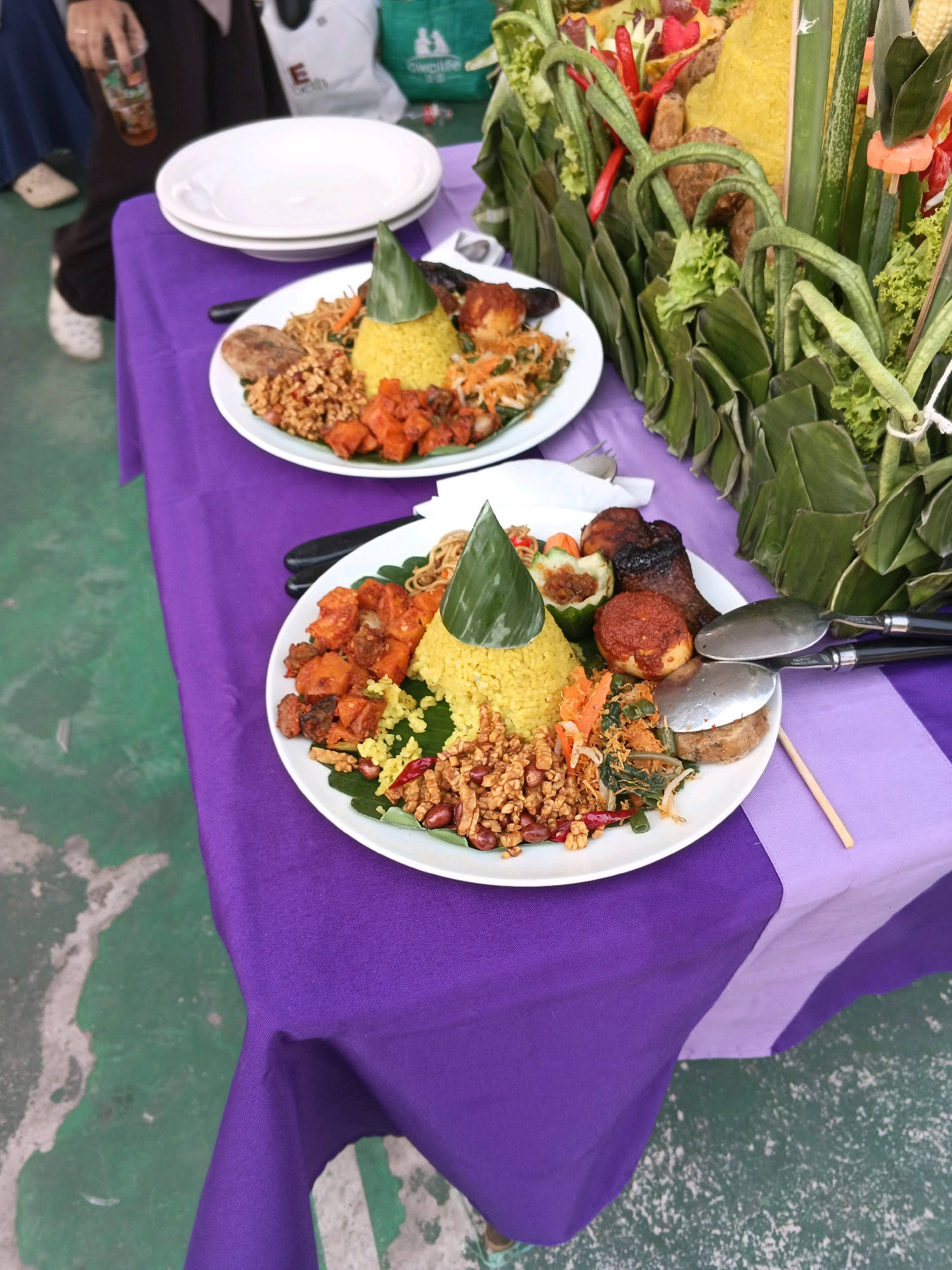 Two plates of Indonesian food, possibly Nasi Tumpeng, are arranged on a purple tablecloth covering a table. The plates contain yellow rice shaped into a cone, surrounded by various side dishes including fried chicken, tempeh, and vegetables. In the background, a large decorative arrangement of food and greenery is visible. A person's hand is holding a drink in a plastic cup. The setting appears to be outdoors, with a green floor visible in the foreground, suggesting a casual event. The table and floor suggest this is likely an outdoor event in a common area, possibly a public space or a backyard gathering. The specific location within Margahayu, Indonesia cannot be determined from the visual information alone.