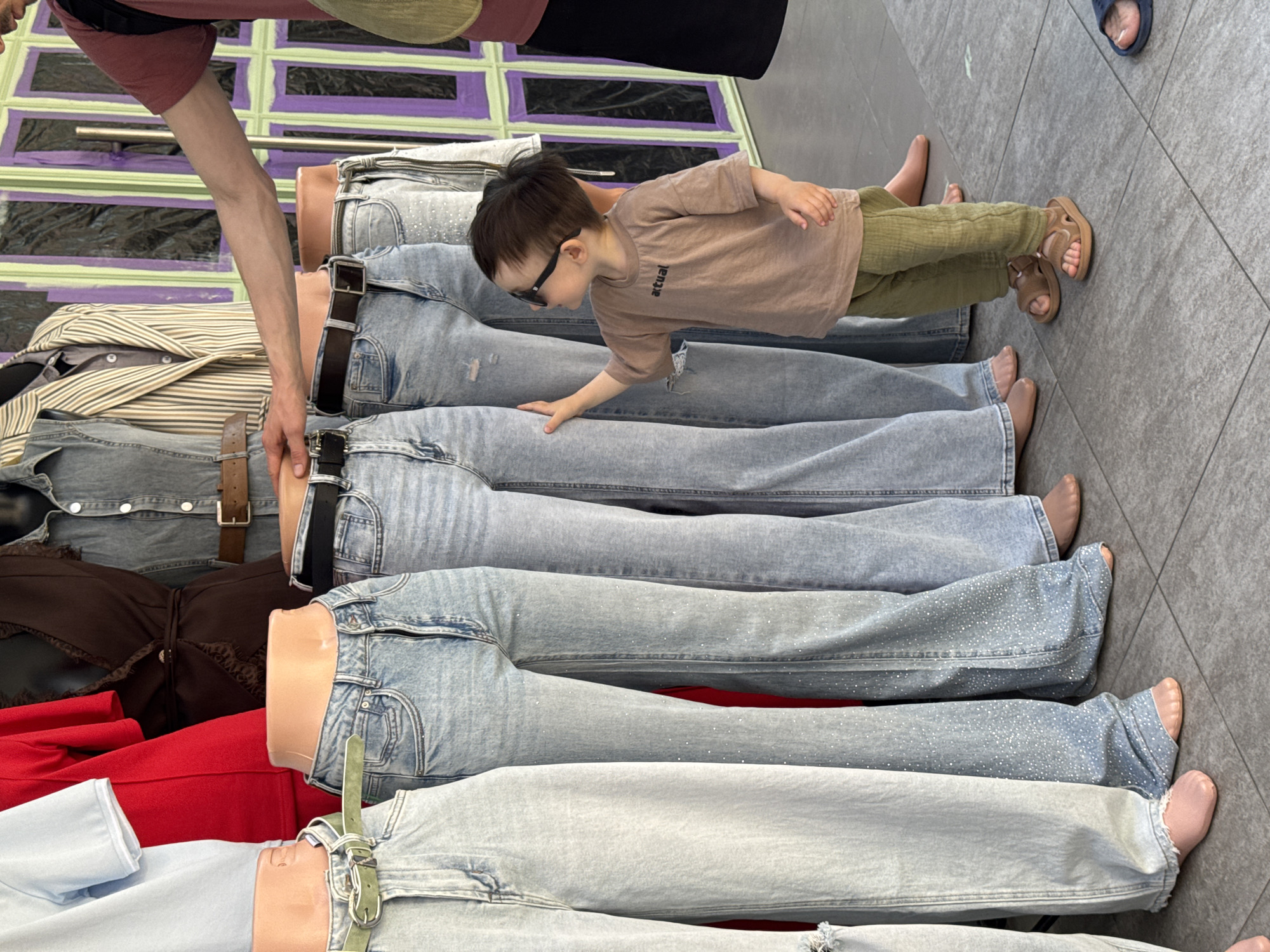 A child wearing sunglasses and sandals is lying on a display of denim jeans worn by mannequins. The mannequins are wearing various tops, including a striped jacket, a brown lace-trimmed shirt, and a red shirt. The child's hand is touching one of the jeans. Another person's arm is also visible in the image. The location could be a retail space in Sochi, Russia.