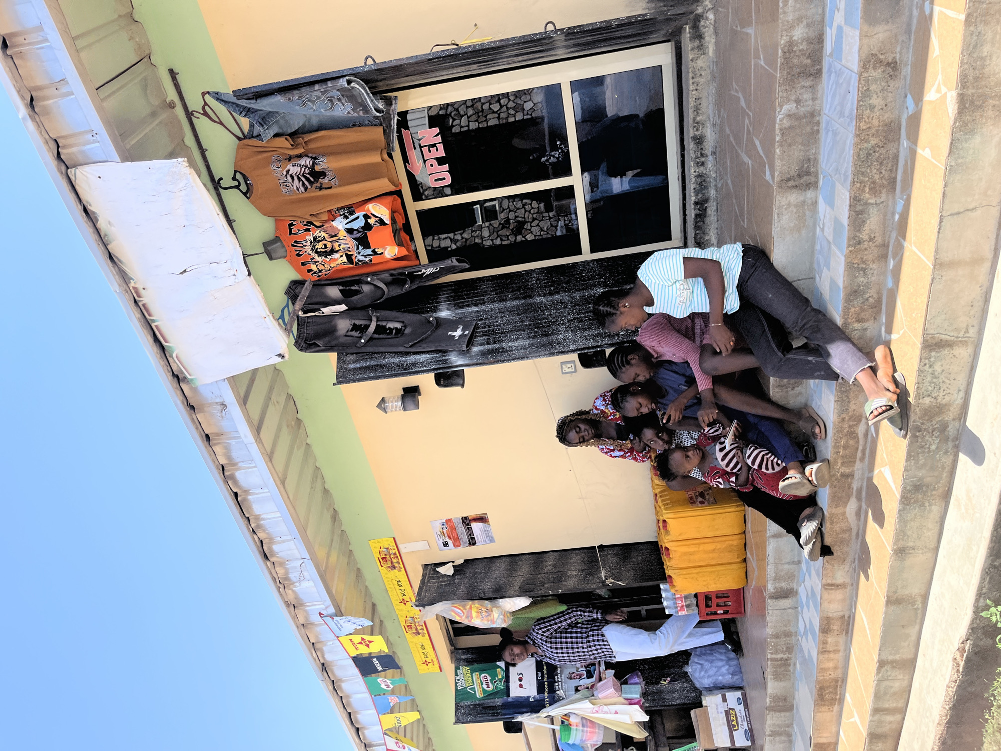 A family of 7 are sitting on the steps of a building in Jos, Nigeria. The building has a storefront with an "OPEN" sign in the window. Clothing hangs on a rack next to the store entrance. The bottom of the building has multiple advertisements. In front of the building are stacked crates and an umbrella is open.