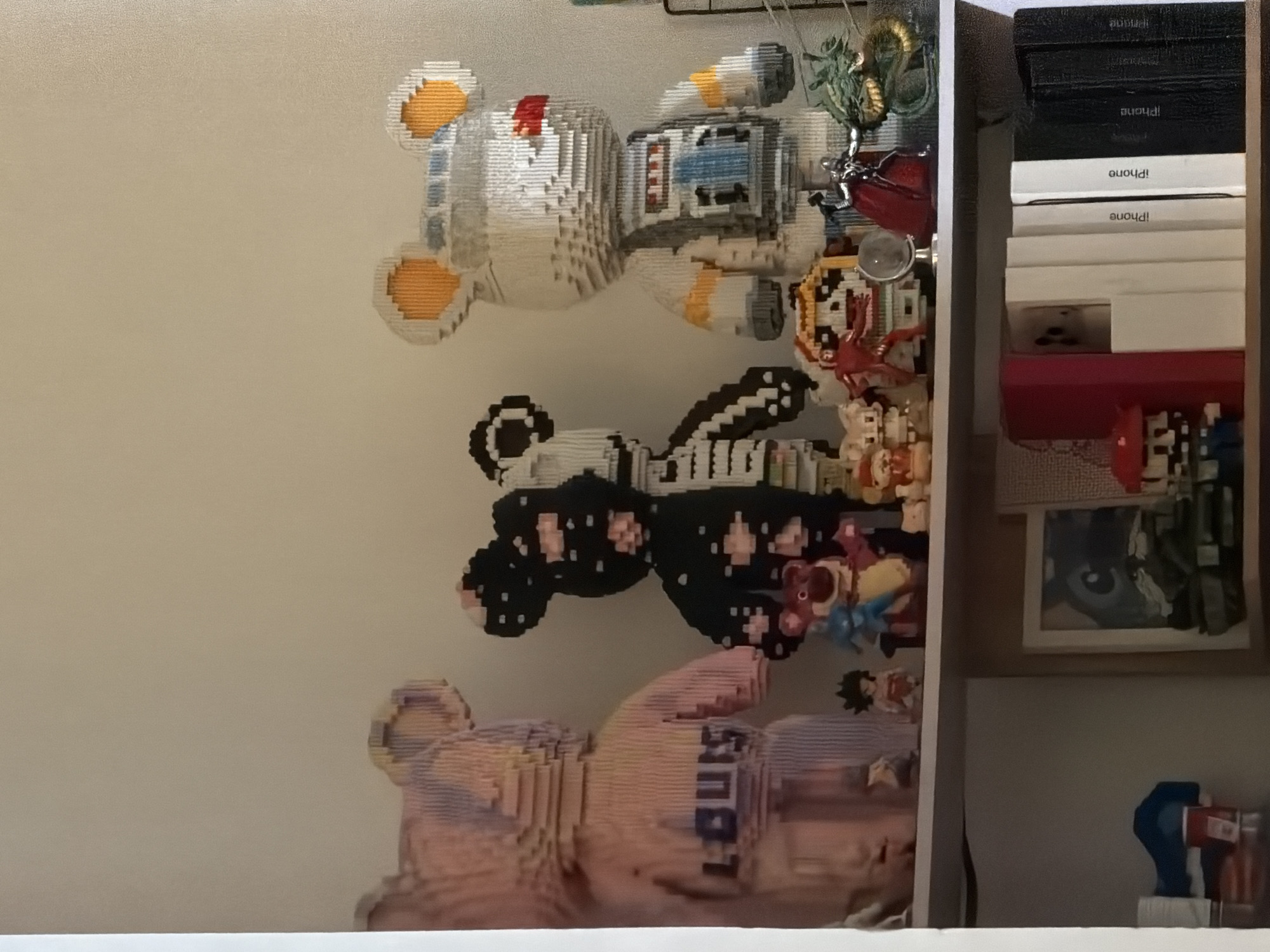 The image shows a shelf with several items. The shelf is white and appears to be a built-in unit. Stacked on the right side of the shelf are several iPhone boxes, alternating between white and black, with the word "iPhone" printed on them. Below the boxes are other items, including a red object and a framed picture with a drawing.
The left side of the shelf features three pixelated bear-like figurines in a vertical arrangement. From top to bottom, they are: a white bear with orange accents, a black bear with white accents, and a pink bear with a possible logo or word on it. The figurines are surrounded by other small toys and decorative items, with some hanging down.
The background is a plain white wall.
There is no information in the image to suggest that the location is in Đà Nẵng, Vietnam.