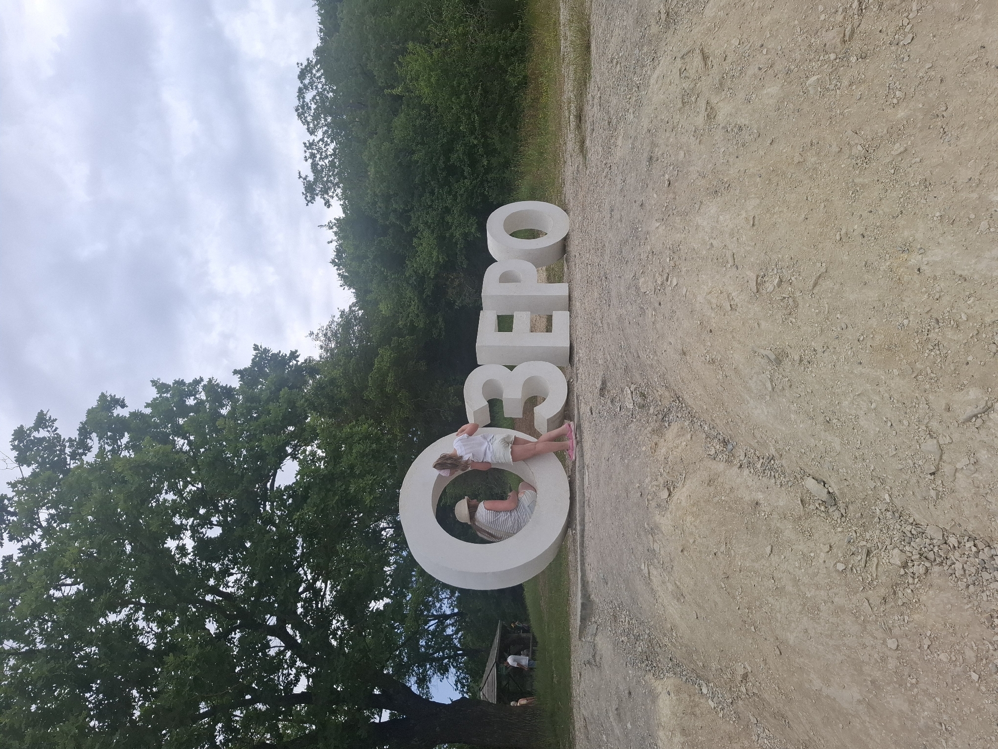 This media file shows a large, white, three-dimensional Cyrillic inscription on a dirt path. The inscription reads "ОЗЕРО," which translates to "LAKE." Two young children are interacting with the letters. One child, wearing a white shirt and shorts, leans against the letter "О," looking into it. Another child, wearing a striped shirt and shorts, is inside the "О," also looking out.
The scene is outdoors, with a backdrop of lush green trees and a cloudy sky. A large oak tree is visible on the left side of the frame. The ground is covered with dirt and small stones, suggesting a natural or park-like setting. In the background, a small wooden structure and other people can be faintly seen, indicating this might be a tourist or recreational area.
The lighting suggests it is daytime, possibly overcast given the sky. The overall mood seems relaxed and playful, with the children enjoying themselves around the large letters. The inscription "ОЗЕРО" and the natural surroundings hint at a location near a lake, likely in Sukko, Russia, as indicated by the location context.