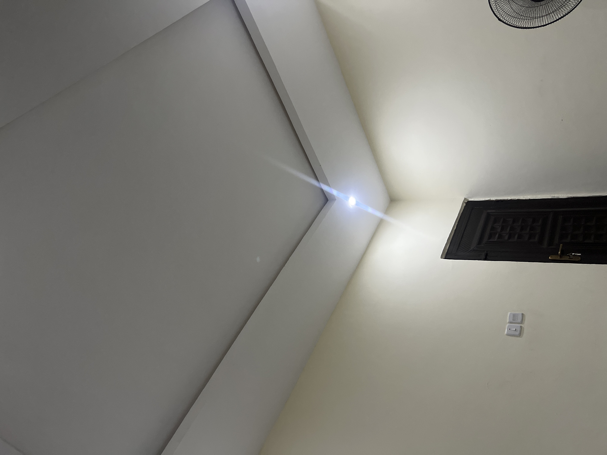 The image depicts an interior corner of a room with cream-colored walls and a white drop ceiling featuring recessed lighting. A dark wooden door with intricate carvings is visible on the right side of the frame, and a portion of a black oscillating fan is present in the far right. On the left wall, two white electrical outlets are mounted. The scene appears to be a residential or office space. Based on the visual cues, this scene could be located in Abuja, Nigeria.