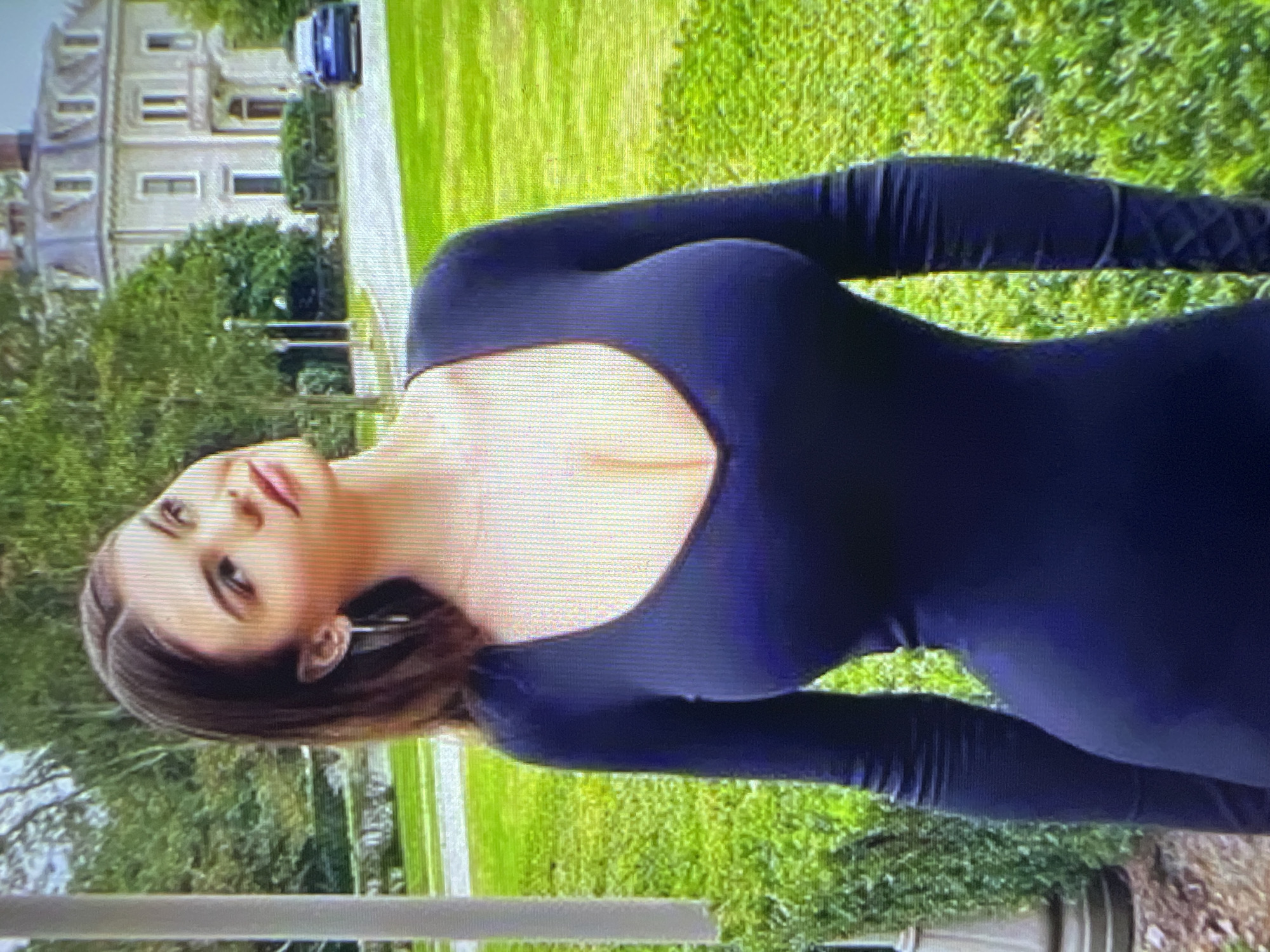A medium shot captures a woman standing outdoors in front of a large, classical-style building. The woman is positioned in the foreground, facing slightly to her right and looking upwards. She has long, dark brown hair that is parted in the middle and falls over her shoulders. She is wearing a dark blue, long-sleeved dress with a V-neckline that reveals a significant portion of her chest. She is also wearing a large, silver hoop earring in her visible ear.
The building behind her is multi-story with a symmetrical facade, multiple windows, and a prominent balcony. Green trees and manicured bushes surround the building, and a well-maintained lawn is visible in the mid-ground. A dark blue car is parked on a paved driveway leading to the building. The overall scene suggests an academic or institutional setting, possibly a university campus. The image is a photograph of a screen, indicated by the visible pixelation and scan lines.