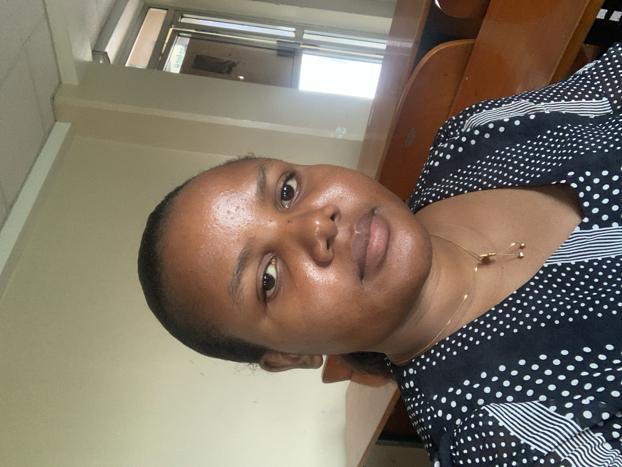 A person is photographed indoors in a bright room, likely a public or semi-public space due to the presence of institutional-style wooden furniture. The individual is a Black woman with dark hair pulled back from her face. She has a gold necklace with small decorative elements. She is wearing a black and white patterned top. The background is light-colored walls and a doorway or window with a metal frame. The location is unknown.
