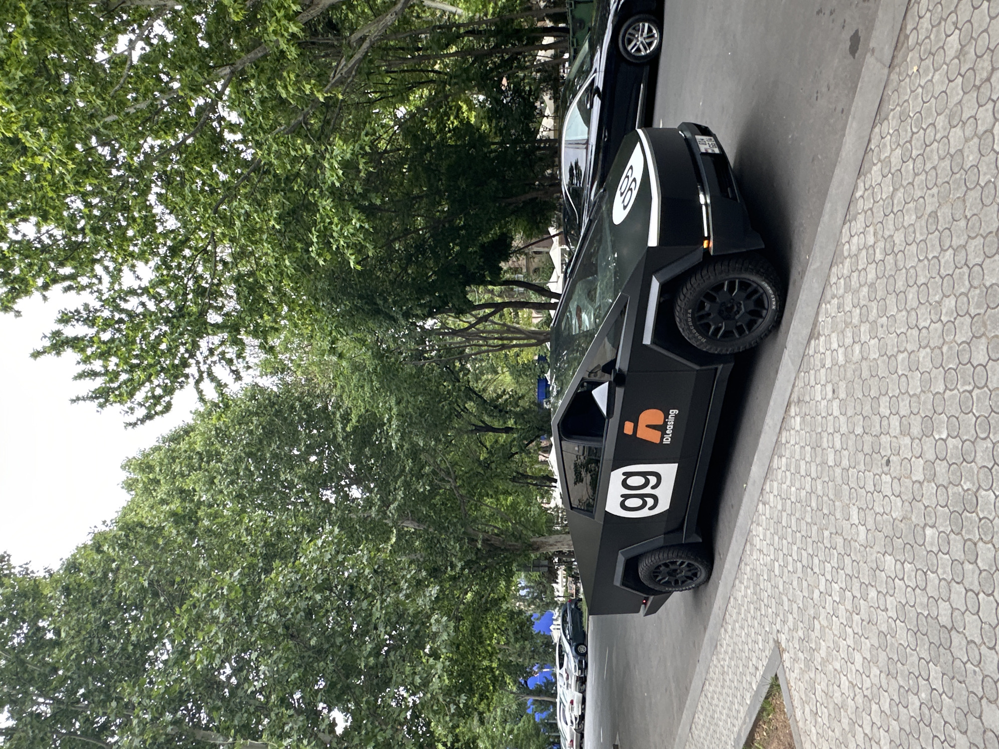 The image depicts a black vehicle with the number "66" prominently displayed on its side, parked on a street in what appears to be Yerevan, Armenia. The vehicle also bears the logo "iDLeasing". The street is lined with trees and other parked cars. A patterned walkway borders the road. The environment suggests an urban setting within the city.