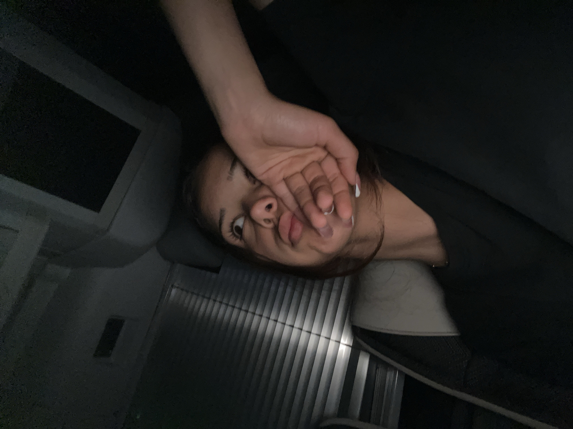 A person is reclining in what appears to be an airline seat. Their right hand is raised, obscuring part of their face. The cabin is dimly lit, and an entertainment screen is visible on the left. The seat appears to be a lie-flat bed type. The city is unknown.