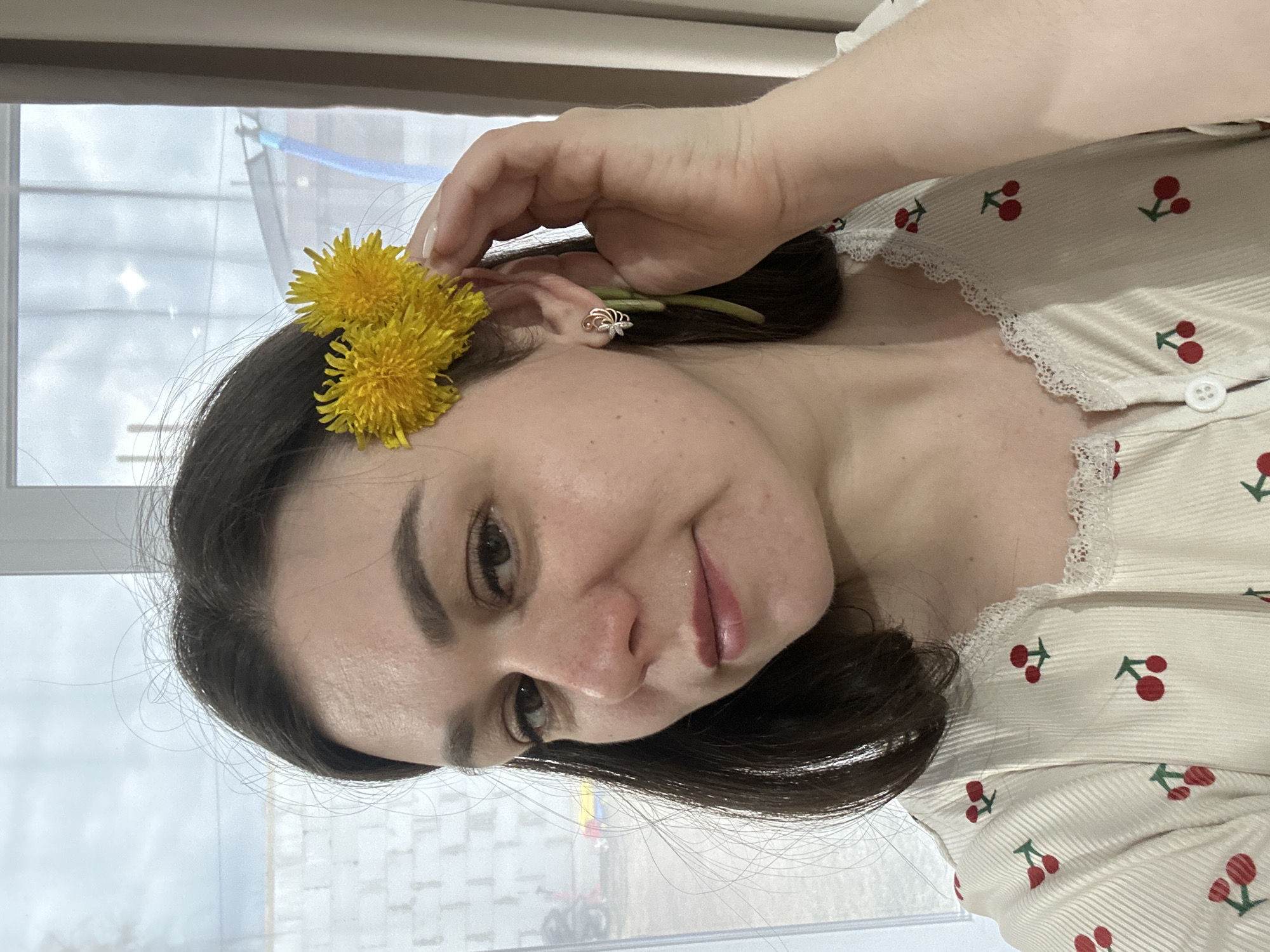 A woman poses indoors. She wears a white top with a cherry pattern and white lace trim. Two yellow dandelions are tucked behind her left ear. Her right hand holds the flower arrangement. The earring is gold. The background features a window with a view of Chaikovsky, Russia. The outside scenery is blurred and unclear.