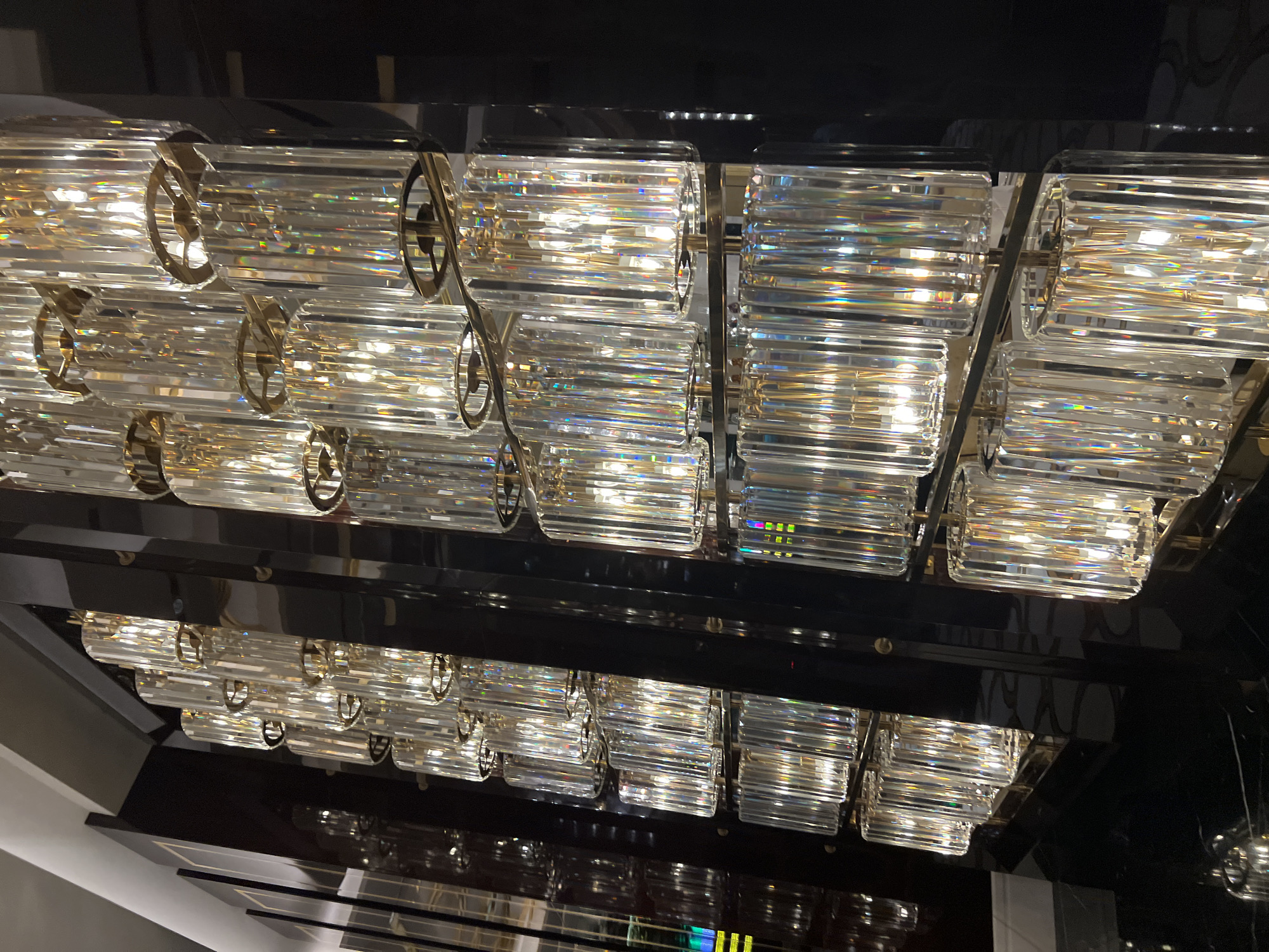 The image shows a close-up, angled view of a modern, multi-tiered chandelier composed of numerous vertical, fluted crystal rods. Each rod is illuminated from within by a warm light source, causing the crystals to refract and scatter light, creating a prismatic effect with rainbow hues. The rods are held in place by horizontal gold-toned metal rings and are arranged in staggered rows on a dark, reflective vertical structure, likely a wall or paneling. The dark surface appears to be polished, possibly lacquered wood or metal, reflecting the light from the chandelier in a distorted manner. The overall impression is one of luxury and opulence, with the interplay of light and crystal dominating the scene. The location is specified as Estosadok, Russia, implying this is an interior decoration within a building in that city. No people are visible in the image.