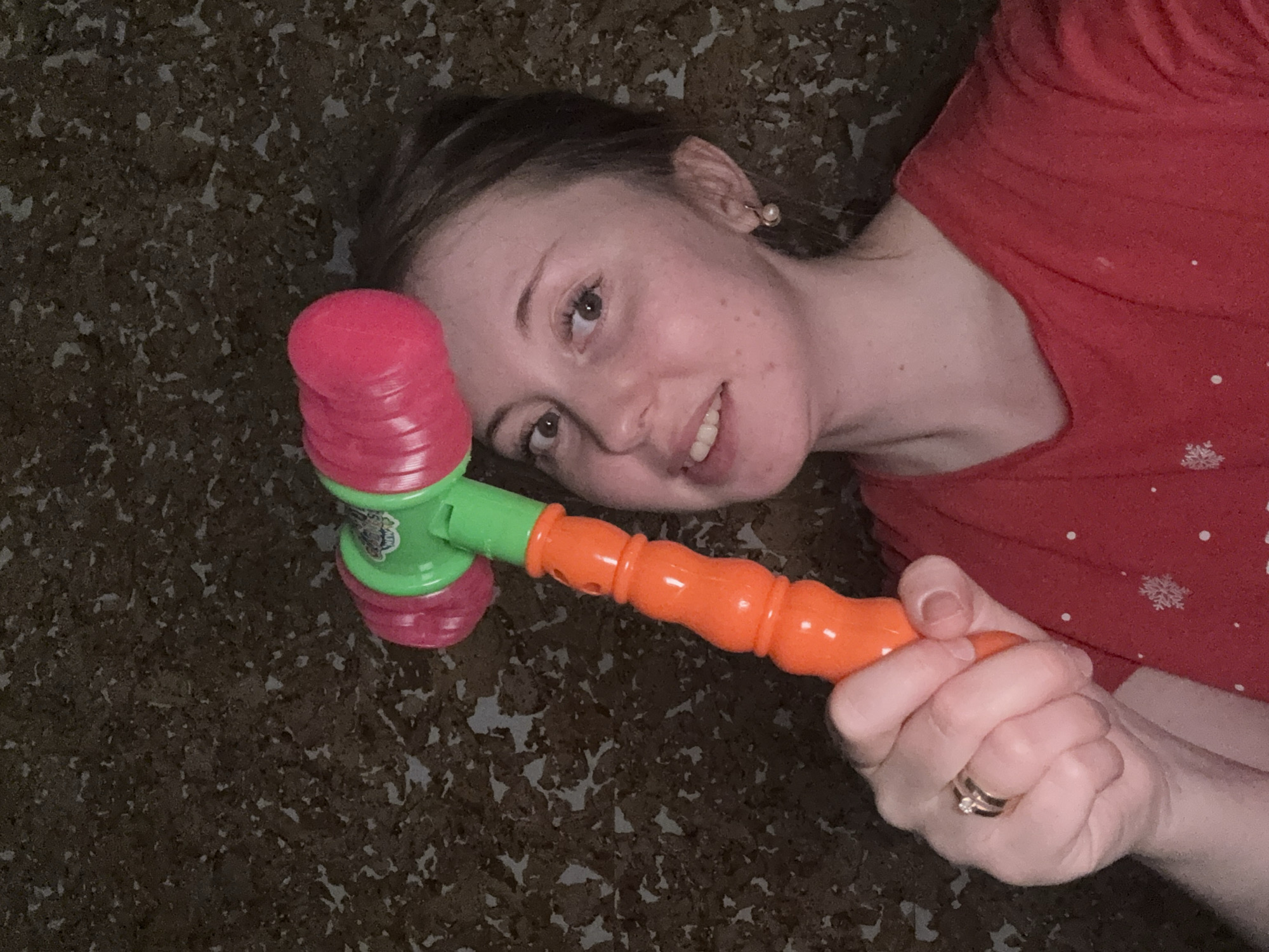 A woman is lying on a surface, possibly a floor, covered in a dark material speckled with light-colored patterns. She is holding a toy hammer with a pink head, green middle section, and orange handle. The woman is wearing a red shirt. She is smiling and looking towards the camera. The location is Unknown, Unknown.