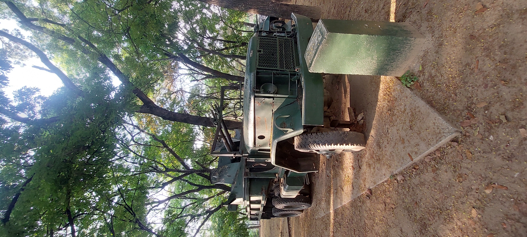 The image displays an olive-green military utility vehicle, possibly a truck or troop carrier, parked outdoors. The vehicle, seen in profile, is rugged in design with a prominent front grille, several tires with visible treads, and a large, box-like extension or attachment on its right side.
The ground beneath and around the vehicle is unpaved, appearing as dirt or earth, with scattered fallen leaves and small debris. A concrete edge or curb is visible in the foreground.
The upper and left portions of the image are dominated by a dense canopy of trees with rich green foliage and visible branches and trunks. Bright sunlight filters through the leaves, casting dappled shadows on the ground and parts of the vehicle, indicating a clear, daytime setting. The bright sky is visible through gaps in the tree cover. No individuals are present in the scene. The setting suggests a natural or park-like area, consistent with locations in Caracas, Venezuela. There is no visible text.