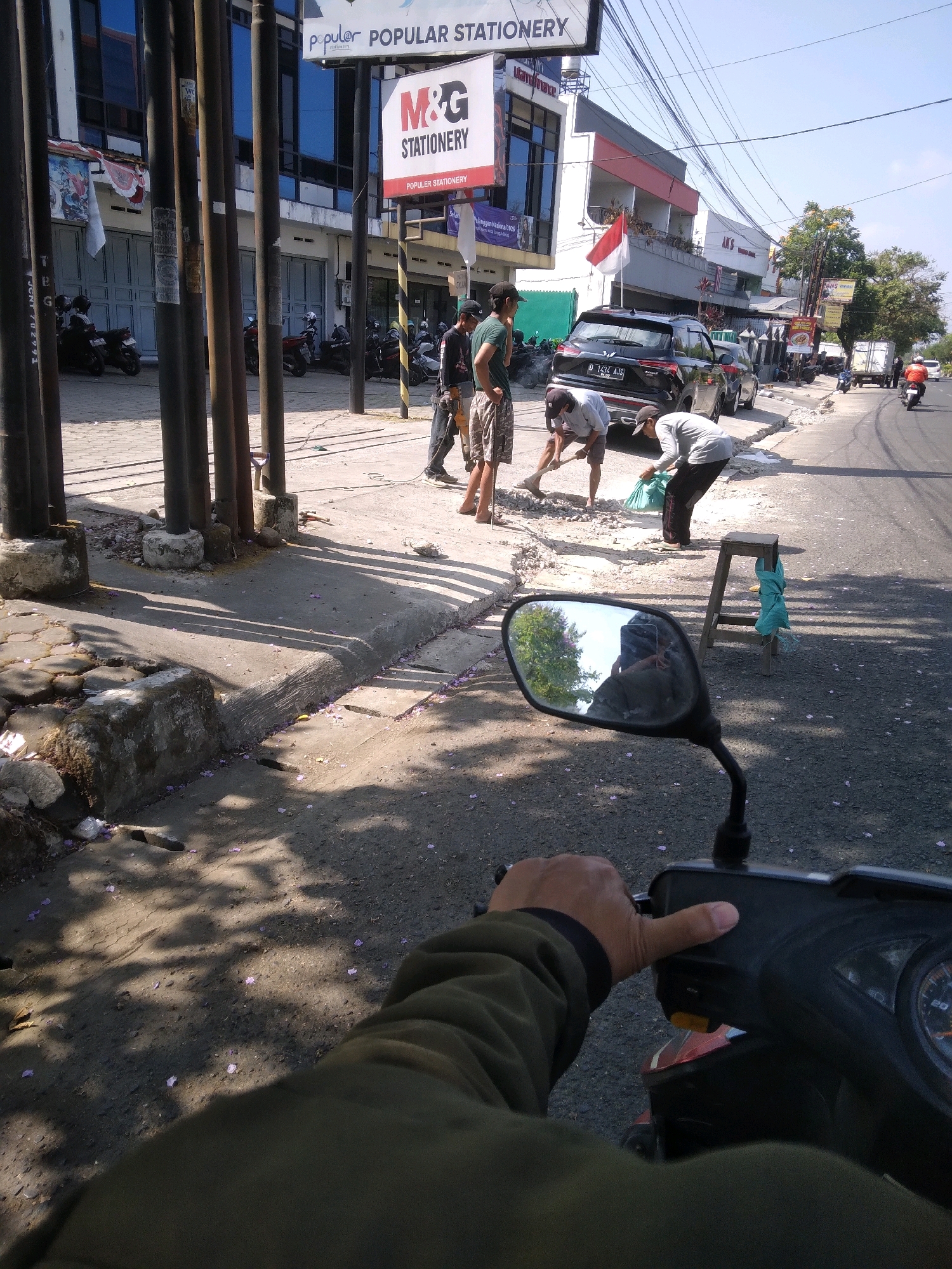 The image captures a scene on a street in Purwokerto Kulon, Indonesia. A person's hand is visible in the foreground, gripping the handlebars of a scooter. In the middle ground, three men are working on the side of the road. Two of them are using shovels to move what appears to be debris or soil, while a third man stands nearby. Behind them, several parked motorcycles and a black car are visible. Buildings line the street, with one prominently featuring a "M&G STATIONERY" sign. The overall environment suggests an active urban street with ongoing work or maintenance.