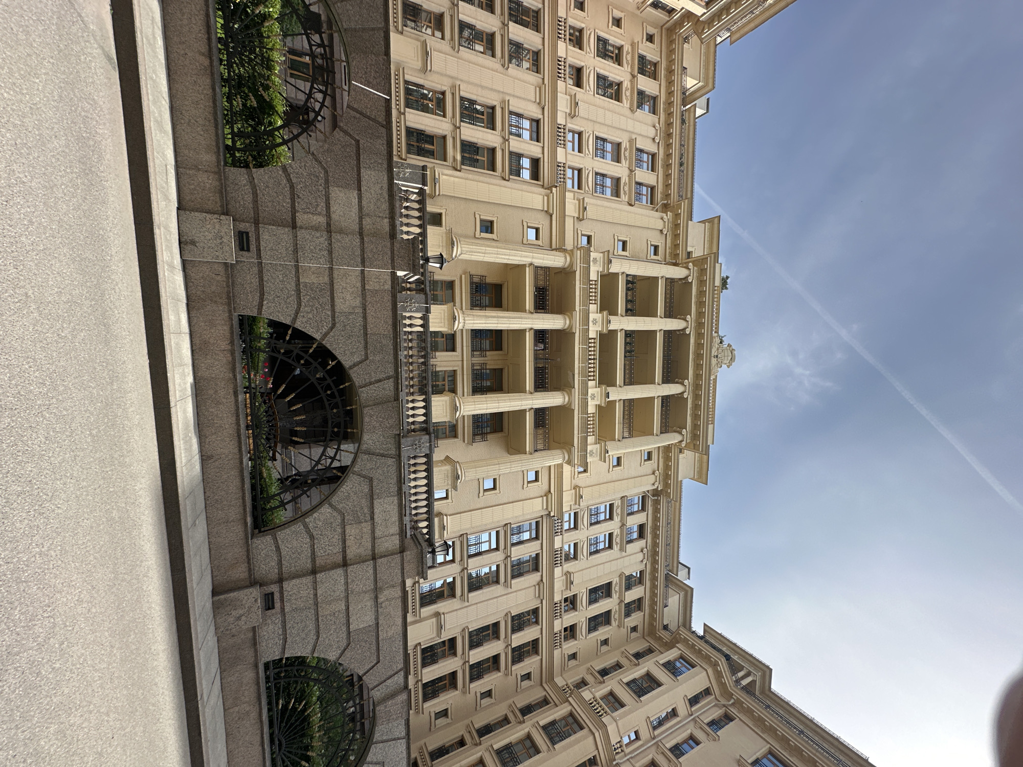 The image depicts a tall, light-colored building with multiple stories in Saint Petersburg, Russia. The building's facade is adorned with architectural details such as columns and balconies. The sky is partially visible with wisps of clouds and a contrail from an airplane. A stone structure with arched openings containing metal grates and greenery is positioned on the left. A concrete sidewalk is on the left side.