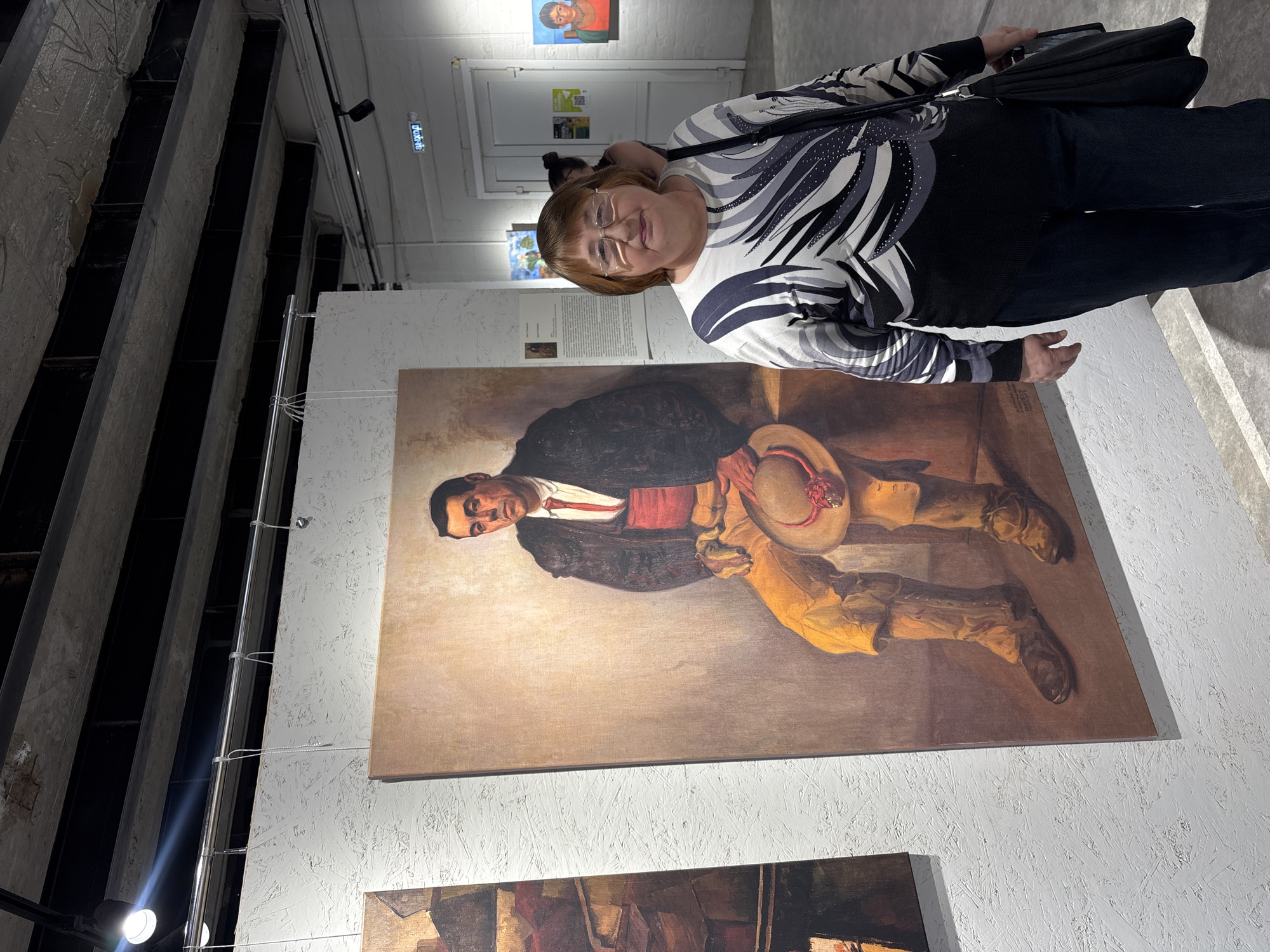 The image shows a woman with short brown hair and glasses standing in what appears to be an art gallery or exhibition space in Krasnoyarsk, Russia. She is wearing a black and white patterned top and dark pants, and carrying a black bag over her shoulder. She is facing towards the painting on the wall. The painting behind her depicts a man in a black jacket, red sash, and yellow pants. He is holding a hat and appears to be seated. Below this painting, another artwork is partially visible. The walls are white with a textured surface. The ceiling has visible structural elements, and there are hanging lights and support cables. In the background, other artwork and signage can be seen, along with other people in the gallery.