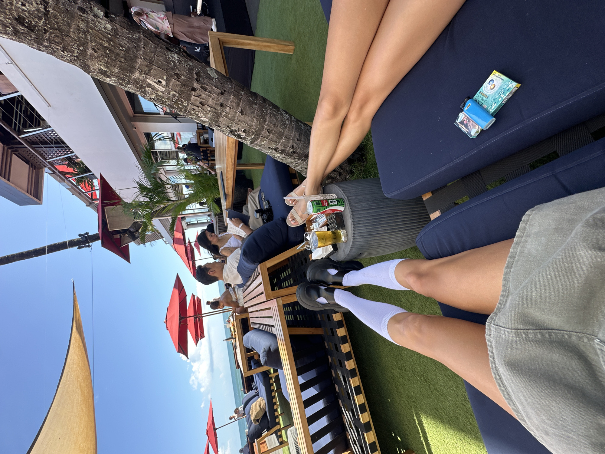 The image is taken at a beachfront establishment in Kuta, Indonesia, likely a bar or lounge due to the presence of beverages and relaxed seating arrangements. A portion of the blue sky and the ocean can be seen in the background. In the foreground are two sets of legs. One set is wearing white socks and black shoes, and the other set is bare with sandals. Cigarettes and a lighter can be seen on the seating next to the bare legs. In the background, people are seated at tables enjoying drinks and food, with red umbrellas providing shade. A large tree trunk is positioned close to the foreground seating, and a portion of a building can be seen in the top left of the frame.