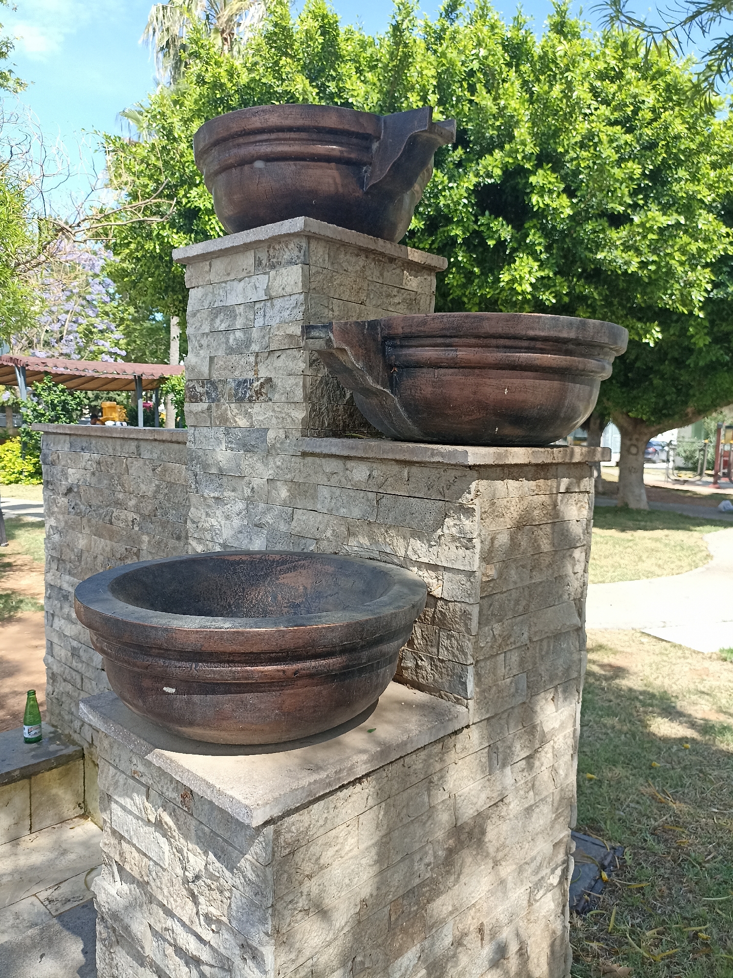 The image shows a tiered fountain in a park-like setting. The fountain is constructed of rough-hewn stone blocks, arranged to create three platforms at different heights. Each platform supports a large, dark-colored bowl that appears to be made of wood or a similar material. The topmost bowl has a notable crack or damaged area on its side. The surrounding environment consists of grass, trees, and a walkway. A green glass bottle is visible on the ground near the base of the fountain. A covered structure with visible chairs or seating is in the background, further indicating a park or public garden. The location appears to be outdoors, with sunlight and clear sky.