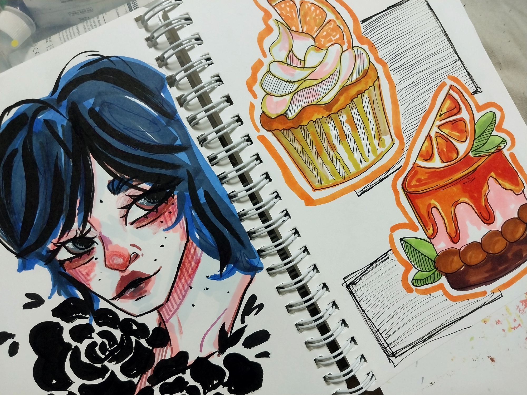 The image shows a drawing in a sketchbook, not a scene in Gimhae-si, South Korea. The drawing features a close-up of a person's face with dark hair, rendered in bold lines and colors, particularly blues, pinks, and black. The person's eyes are large and expressive. To the left of the face, there is a black, abstract floral or leafy design. Below the face, occupying the lower half of the sketchbook page, are two drawings of food items outlined in orange marker. One is a slice of cake with yellow and white striped layers and pink frosting topped with a slice of orange. The other is a rectangular piece of food, possibly a cake or pastry, with brown circular elements resembling berries and two green leaves, also adorned with an orange slice. Both food items are placed on a surface depicted with diagonal black lines within rectangular outlines. The sketchbook is open, and a spiral binding is visible along the top edge of the lower section.