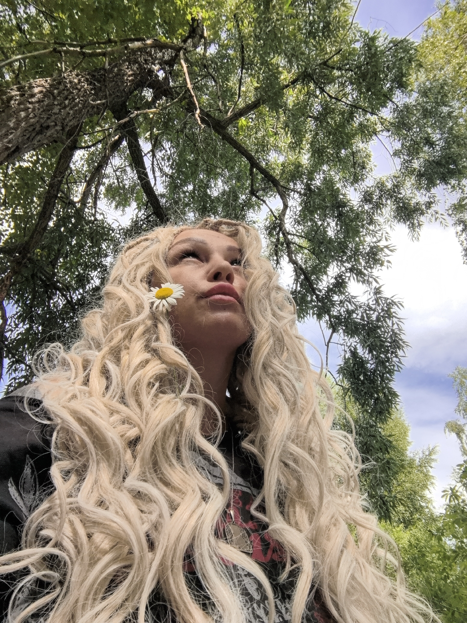 The image shows a low-angle, close-up shot of a young woman with blonde, curly hair and a daisy tucked behind her ear. She is looking upwards, with her lips slightly parted. She is wearing a dark t-shirt with a graphic design. The background consists of green trees with sunlight filtering through the leaves, and a blue sky with some clouds. The perspective suggests the photo was taken outdoors, possibly in a park or wooded area. There is no specific indication of the location being Noviy, Russia.