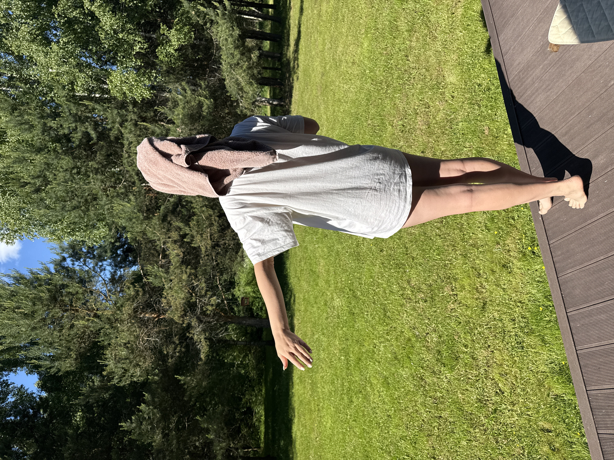 An outdoor, sunlit scene in Копнино, Russia, depicts one individual from behind, standing barefoot on a dark brown slatted wooden deck. The person, whose gender is not explicitly identifiable but appears to be female, is wearing a loose-fitting white t-shirt that extends to mid-thigh. A light brown towel is draped over their head and shoulders, partially obscuring the head. Their right arm is extended straight out to the side, palm open and facing forward, with fingers slightly spread and nails painted white. A distinct shadow of the person is cast on the deck, indicating bright sunlight from the left. Beyond the deck, a large expanse of vibrant green grass, interspersed with small yellow flowers, fills the midground. The background consists of a dense forest line dominated by tall pine trees and some lighter-barked deciduous trees. Patches of clear blue sky with minimal white clouds are visible through the tree canopy. A small corner of a light-colored cushion or mattress is visible on the far right of the wooden deck.