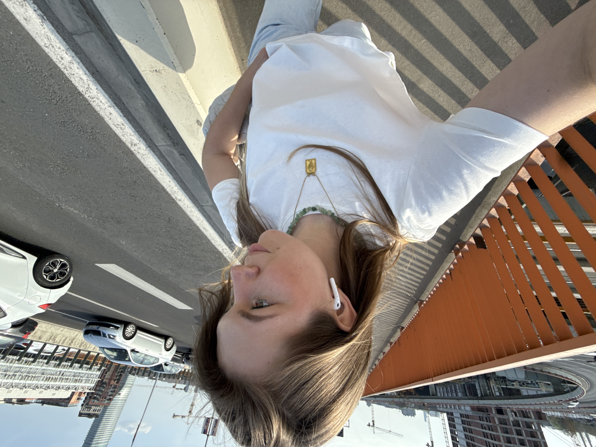 This image, captured on an elevated bridge or overpass, features a young woman as the primary subject, likely taking a selfie. She is shown from the chest up, with light brown hair, wearing a plain white t-shirt, light-colored pants, and a white earbud in her right ear. Two necklaces are visible: one with a rectangular golden pendant and another crafted from green beads.
The setting is an urban environment, identified as Belgrade, Serbia. To the woman's right, a multi-lane road with visible cars, including a white hatchback and a silver sedan, spans the foreground. An orange metal railing or fence runs alongside the bridge, casting prominent, striped shadows across the ground and the woman's lower body, indicating bright, direct sunlight.
In the background, a sprawling city skyline is visible under a clear sky. Modern buildings, including a tall, slender skyscraper and various other structures, punctuate the landscape. Numerous construction cranes are scattered throughout the background, signaling active urban development. The time of day is daytime, characterized by strong sunlight and distinct shadows. The woman's expression appears neutral and composed. No visible text is present in the image.