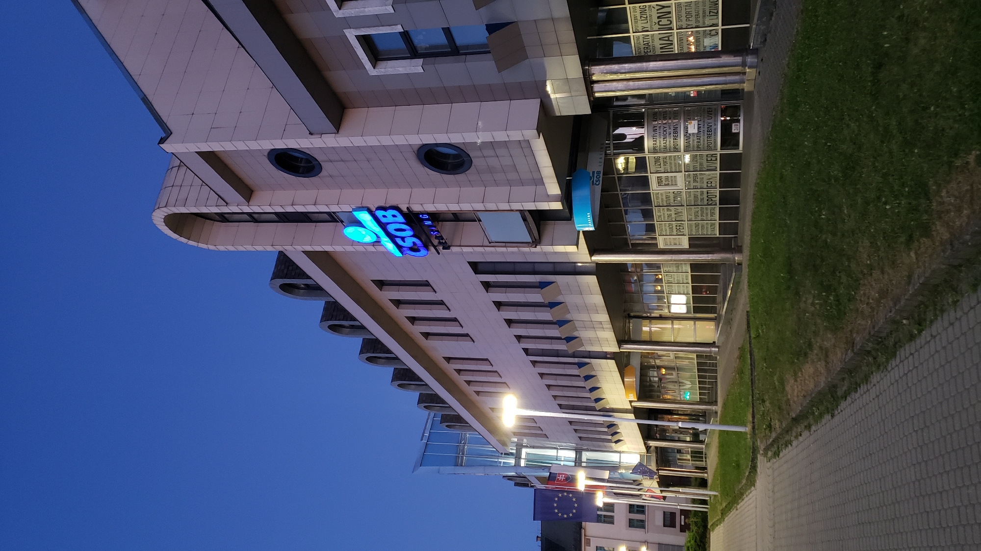 The image depicts the exterior of a modern commercial building at dusk in Prešov, Slovakia. The building features a light-colored facade with a distinctive arched roofline and multiple windows. A prominent blue neon sign reading "ČSOB LEASING" is affixed to the upper portion of the building. Several flags, including the European Union flag and the Slovak flag, are visible on poles to the left of the building. The foreground shows a paved walkway and a grassy area. The sky is a deep blue, indicating early evening. There are no people visible in the image. The overall scene is one of urban commercial architecture under twilight conditions.