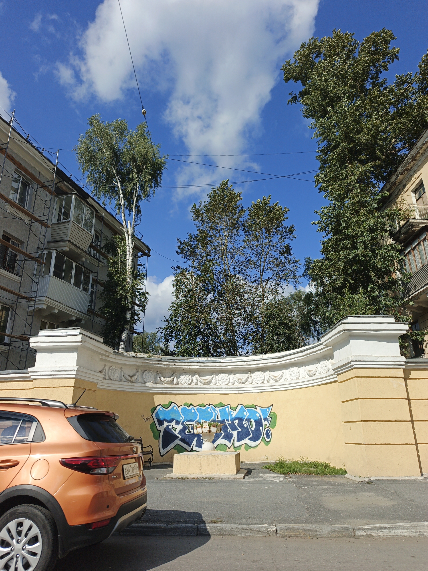 In Zlatoust, Russia, on a sunny day with a blue sky and scattered clouds, an orange Kia Rio is parked on the right side of the street. The car is facing away from the camera and to the left, with its rear visible. The car's license plate is partially obscured but appears to be E647XP 174.
Adjacent to the street is a yellow wall with ornate white decorative elements along its top. On the wall, there is graffiti featuring blue and green stylized lettering. To the left of the graffiti, a concrete pedestal is visible, and behind it, a small patch of green grass.
Behind the wall, taller trees with green foliage are prominent. To the left, a multi-story building with balconies is visible, partially covered by scaffolding. The building appears to be undergoing renovation. The overall scene depicts an urban environment with elements of construction, street art, and nature.
