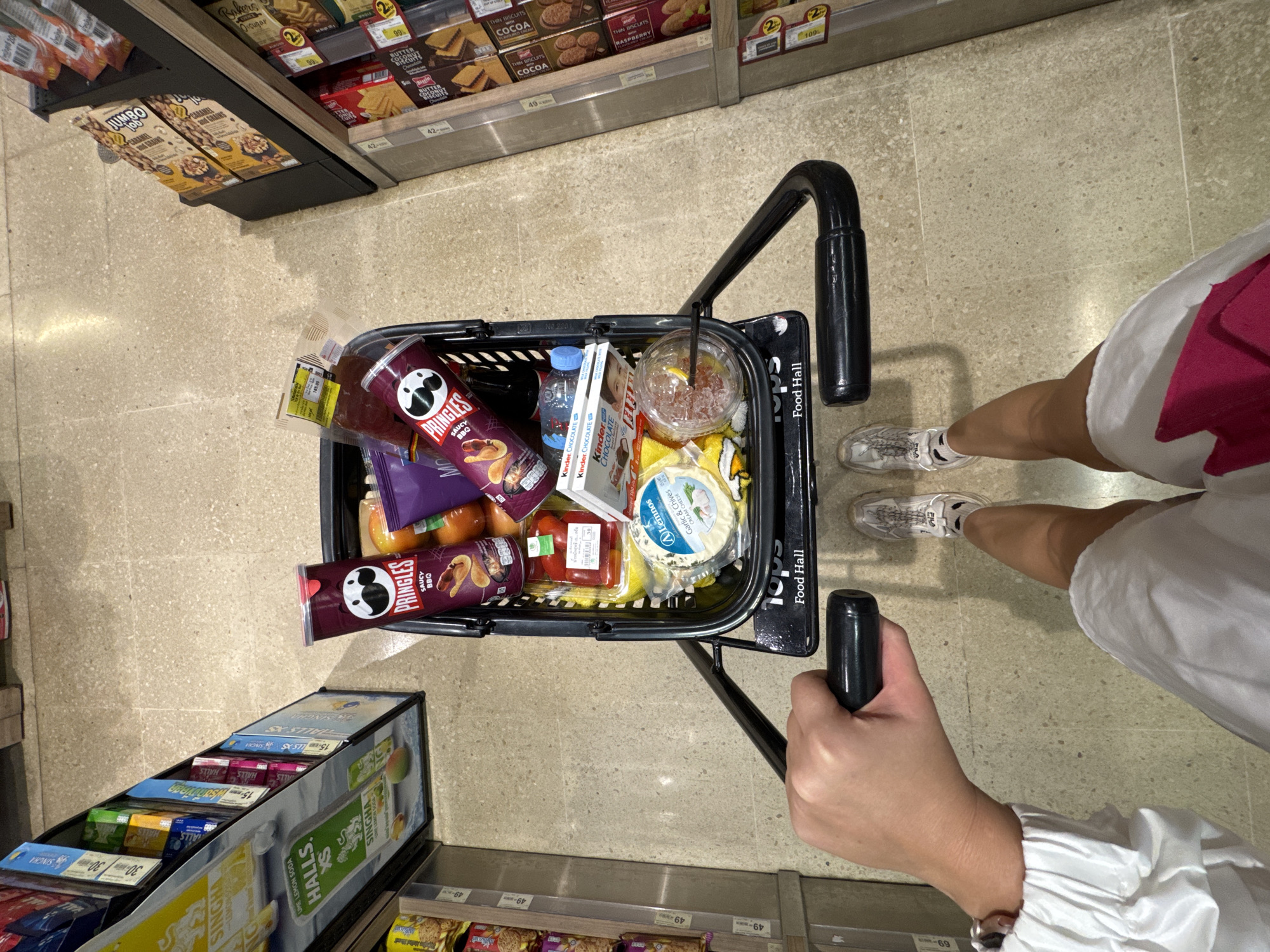 The image shows a shopping cart filled with groceries, viewed from directly above. The cart contains two cans of Pringles, a bottle of water, two boxes of Kinder Chocolate, a cup with a straw, a container of cheese, a package of tomatoes, and other items. The handle of the cart is being held by a hand with a white sleeve, while the legs of the person holding the cart are visible, wearing white shorts with a pink stripe and grey sneakers. The cart is in a supermarket aisle, with shelves of products visible on both sides. The products on the shelves include biscuits, crackers, candies, and other snacks. Price tags are visible on the shelves. The cart has "tops Food Hall" printed on its side. The OCR confirms that some products are SINGHA Lemon Soda and HALLS candies. Given that Food Hall is part of Central Group, the location is most likely in a Central Group affiliated retail space, or supermarket in Chiang Mai City Municipality, Thailand.