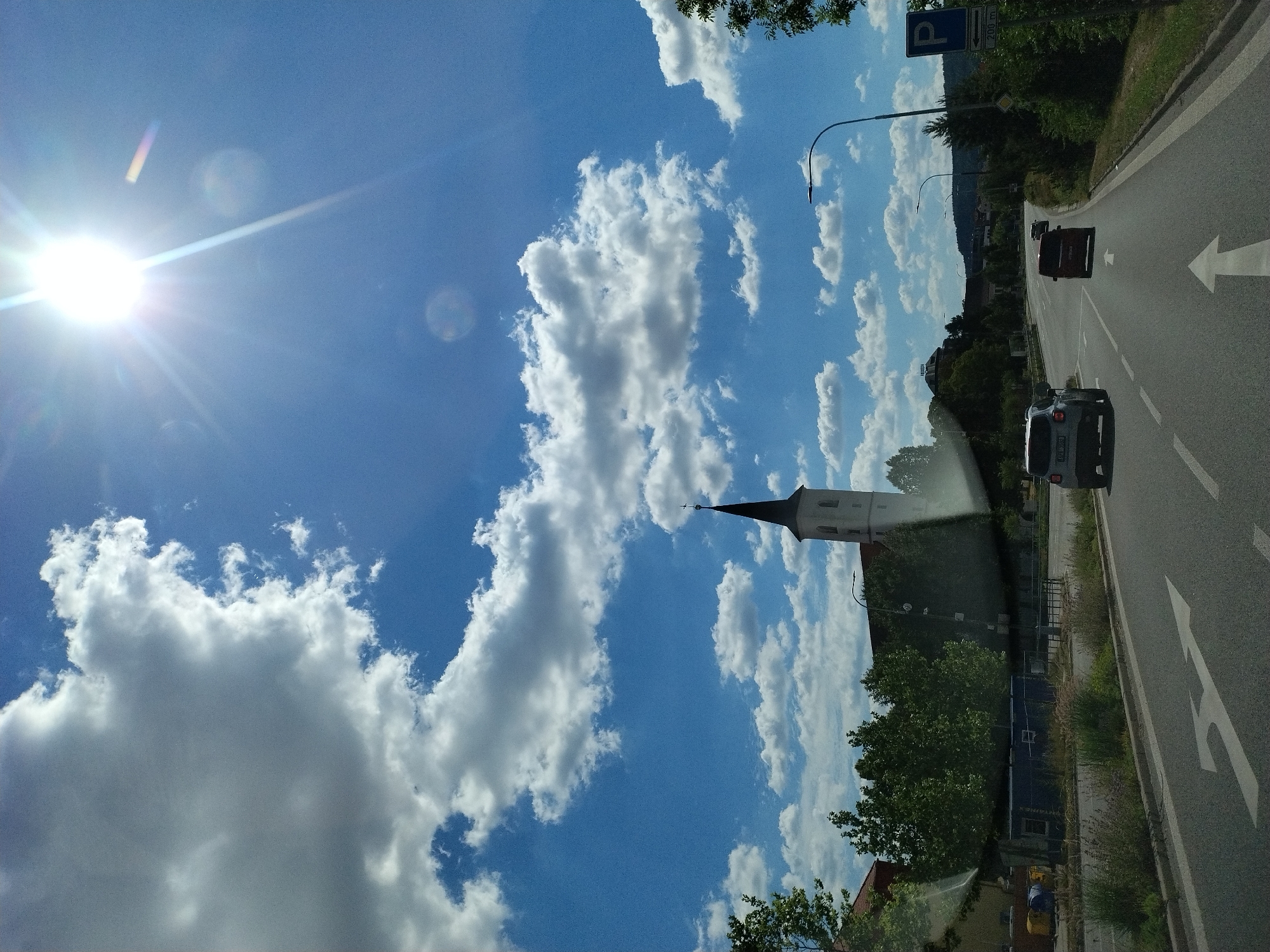 This is a photograph taken from a moving vehicle, looking out through the windshield. The scene depicts a road in Strakonice, Czechia, on a sunny day with scattered white clouds in a bright blue sky. The sun is high and bright, creating lens flares and starburst effects in the image.
In the foreground, a two-lane asphalt road is visible, with white lane markings. The vehicle from which the photo is taken is traveling on the right lane, with a white arrow painted on the road indicating a choice of going straight or turning right. Two cars are ahead: a white SUV in the right lane and a dark red minivan in the left lane. Further down the road, a smaller, lighter-colored car is visible in the distance.
To the left of the road, there are some buildings and trees. A prominent white church with a dark spire and a cross on top stands against the sky, surrounded by green trees. In front of the church, there are some temporary blue structures and yellow bins.
To the right of the road, there is a sidewalk or verge with vegetation, followed by trees and then a blue parking sign indicating a parking area 200 meters ahead. Beyond the immediate roadside, the landscape appears to be hilly with more trees and buildings visible in the distance. The overall impression is a suburban or semi-rural setting.