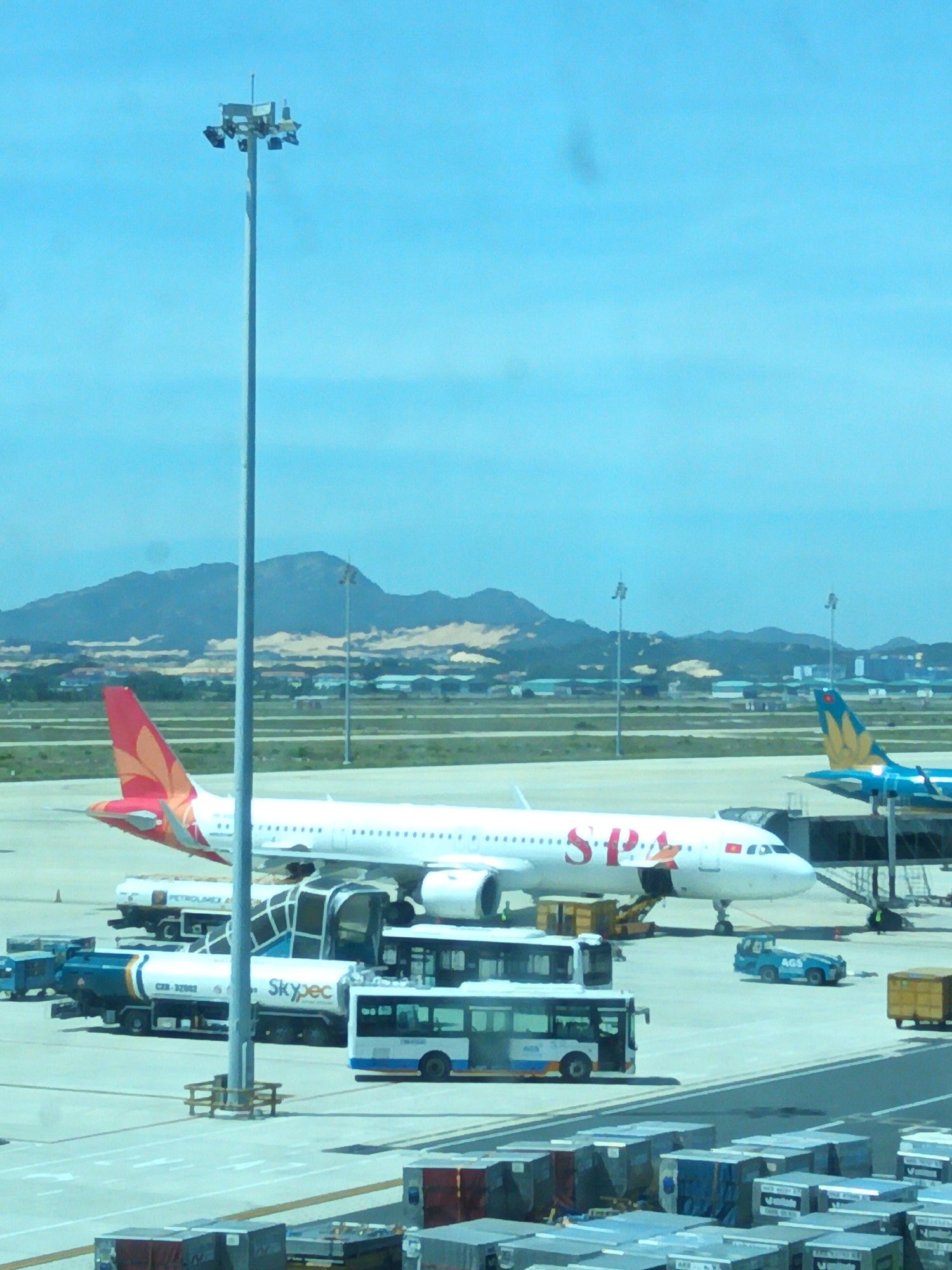 The image captures an outdoor scene at an airport in Bắc Cam Ranh, Vietnam, under a bright, clear blue sky. In the foreground, several stacks of cargo containers are visible on the tarmac. A tall light pole with multiple lamps stands prominently in the center-left of the frame.
Dominating the midground are two airplanes. The airplane on the left is predominantly white with a red and orange tail fin featuring a floral design. The text "SPA" is visible on its fuselage. Adjacent to this aircraft is a fuel truck with "Skypec" branding. A bus is also parked near the fuel truck. Further to the right, the tail of a second airplane, painted in blue with gold accents, is visible.
In the background, a range of mountains forms the horizon, with scattered buildings and infrastructure suggesting an urban or semi-urban landscape. The overall impression is of a busy airport environment during daylight hours.
