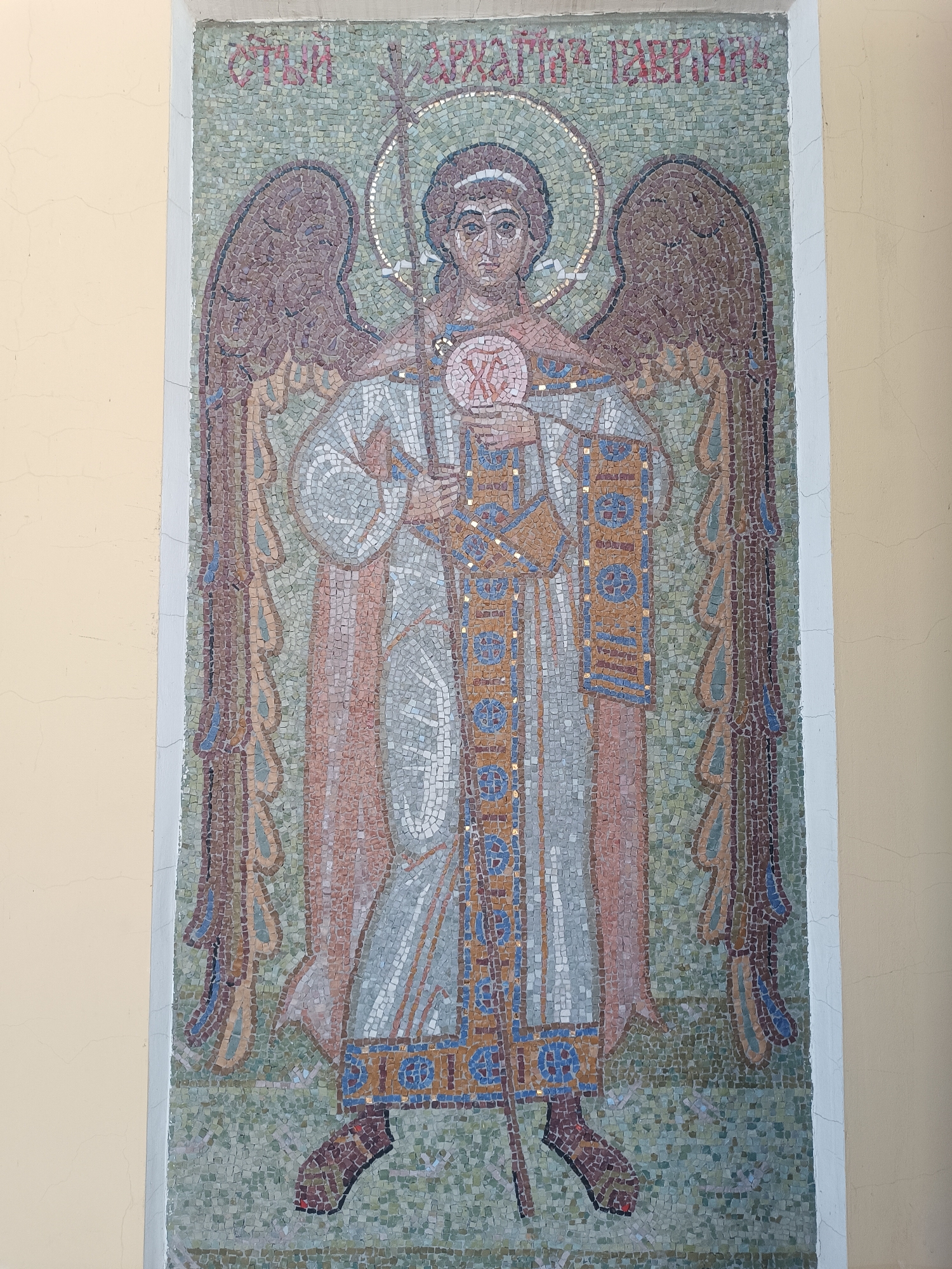 The image shows a mosaic depicting an angel with wings, holding a spear and an orb with Cyrillic characters. The mosaic is set against a pale yellow wall with some minor cracks and imperfections. Above the figure is Cyrillic text. This mosaic is located in Moscow, Russia.