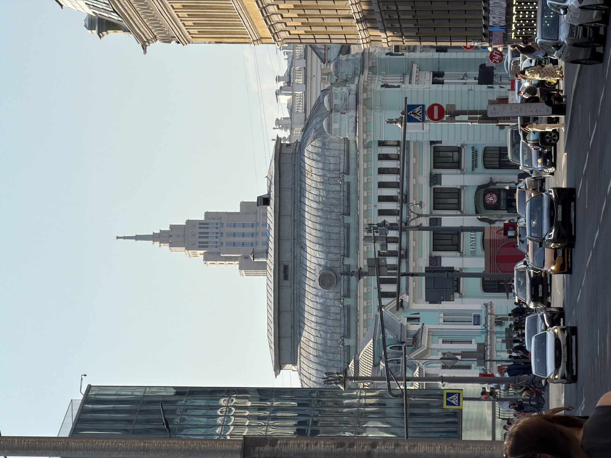 This photograph captures a street scene in Moscow, Russia, featuring a combination of architectural styles and modern urban life. The foreground includes a modern glass building and elements of street infrastructure like traffic lights and signs. Moving into the midground, there are a number of parked cars along the right side of the street, with people walking and interacting on the sidewalk beside the cars. On the left side of the street stands a row of buildings with classical architectural details, including one prominent building with a distinctive curved roof. In the background, one can make out a skyscraper. The sky is clear.