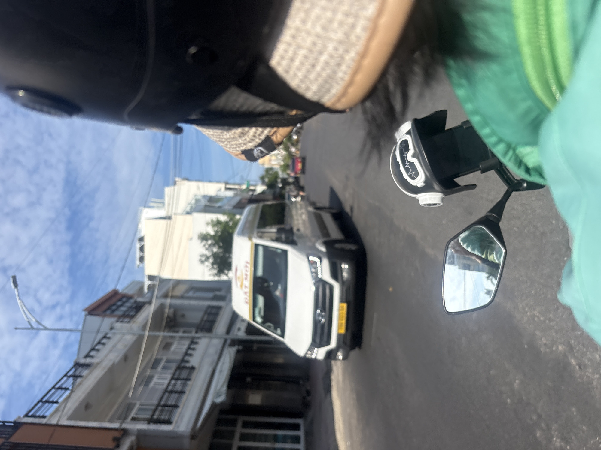 The image captures a street scene in Nha Trang, Vietnam, from the perspective of someone riding a motorcycle. In the foreground, a portion of the rider's helmet and a green jacket are visible. A small, decorative helmet is mounted on the handlebars. To the left, a motorcycle's side mirror reflects the road and buildings.
The main subject on the road is a white van, likely a passenger van, with "ĐẠT MỚI" written on its top front. The van has a yellow license plate. Further down the street, other vehicles are present, including a red car. The environment features buildings lining the street, with balconies and architectural details typical of urban Southeast Asia. The sky is blue with scattered clouds, indicating daylight conditions. The road surface is asphalt. The overall impression is a snapshot of daily life in a Vietnamese city, with traffic and urban infrastructure as the backdrop.