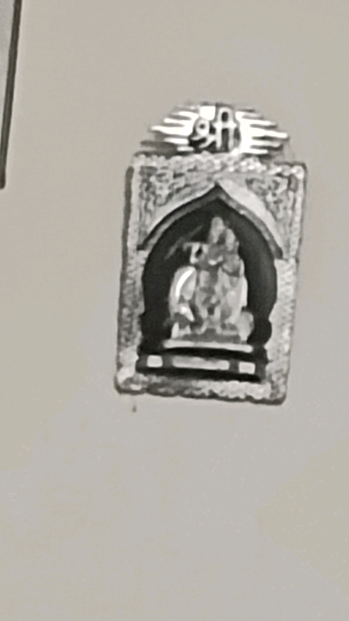 The image shows a close-up of a silver pendant or amulet. At the top of the object, there are stylized markings that appear to be the Hindi word "Shri". Below this, the object is rectangular with a textured, embossed border. Within this border, there is an arched recess containing a small, detailed sculpture of a deity, likely Hindu. The deity is depicted seated, with multiple arms. The overall style suggests religious or devotional significance. The background is a plain, light-colored surface, likely a wall or tabletop, providing no contextual information about its specific location within the city of Deoghar, India.