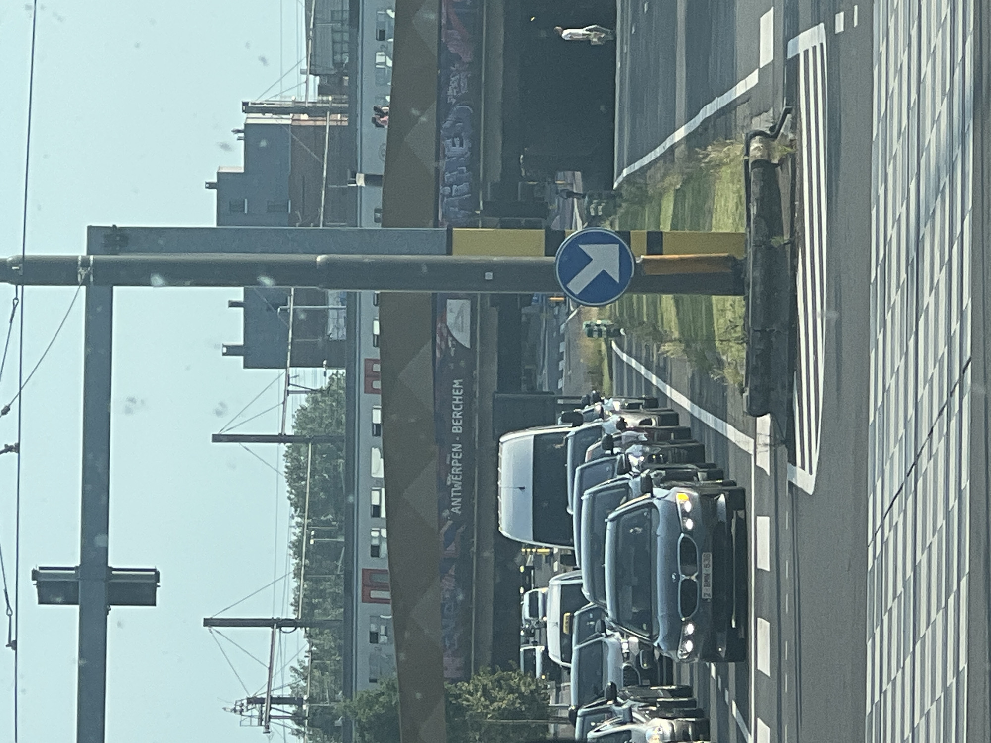 The image shows a traffic jam on a road in Antwerp, Belgium. A line of cars, including a dark BMW with license plate "Z-BMW-638", is stopped at a crosswalk. In the background, an elevated train line with graffiti on it crosses the road. The sky is clear and blue.