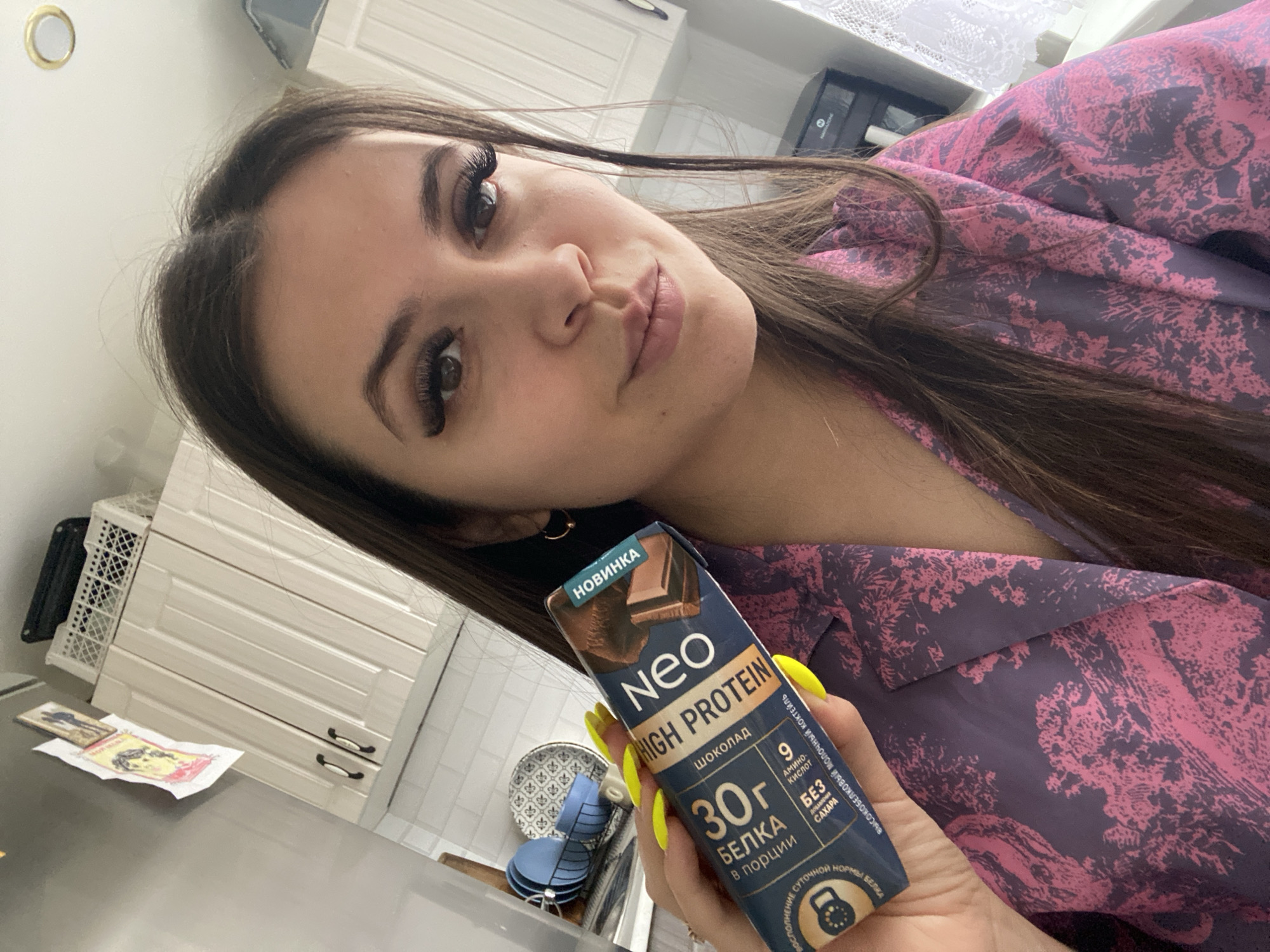 A woman is holding a protein shake by NEO high protein chocolate. The protein shake has Russian text indicating it has 30g of protein per serving. The location is indoors, likely a kitchen, with white cabinets in the background. The woman is wearing a pink and grey patterned top. Her fingernails are painted yellow. The protein shake box states that the product is made in Chaikovsky, Russia.
