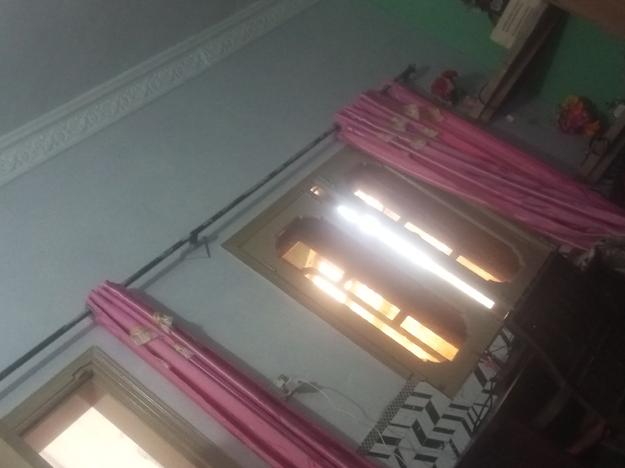 The image shows an interior scene, possibly a room. There are pink curtains visible, with some decorative elements. One window or opening has a light source shining through it, creating bright rectangular shapes. The wall appears to have decorative molding near the ceiling. In the background, there is a dark object that could be furniture or a shelf, and some decorative items that look like flowers. The floor has a patterned tile design. The overall impression is that of a residential or a similarly private indoor space, likely located in Mansehra, Pakistan, given the context. There are no people or specific actions depicted.