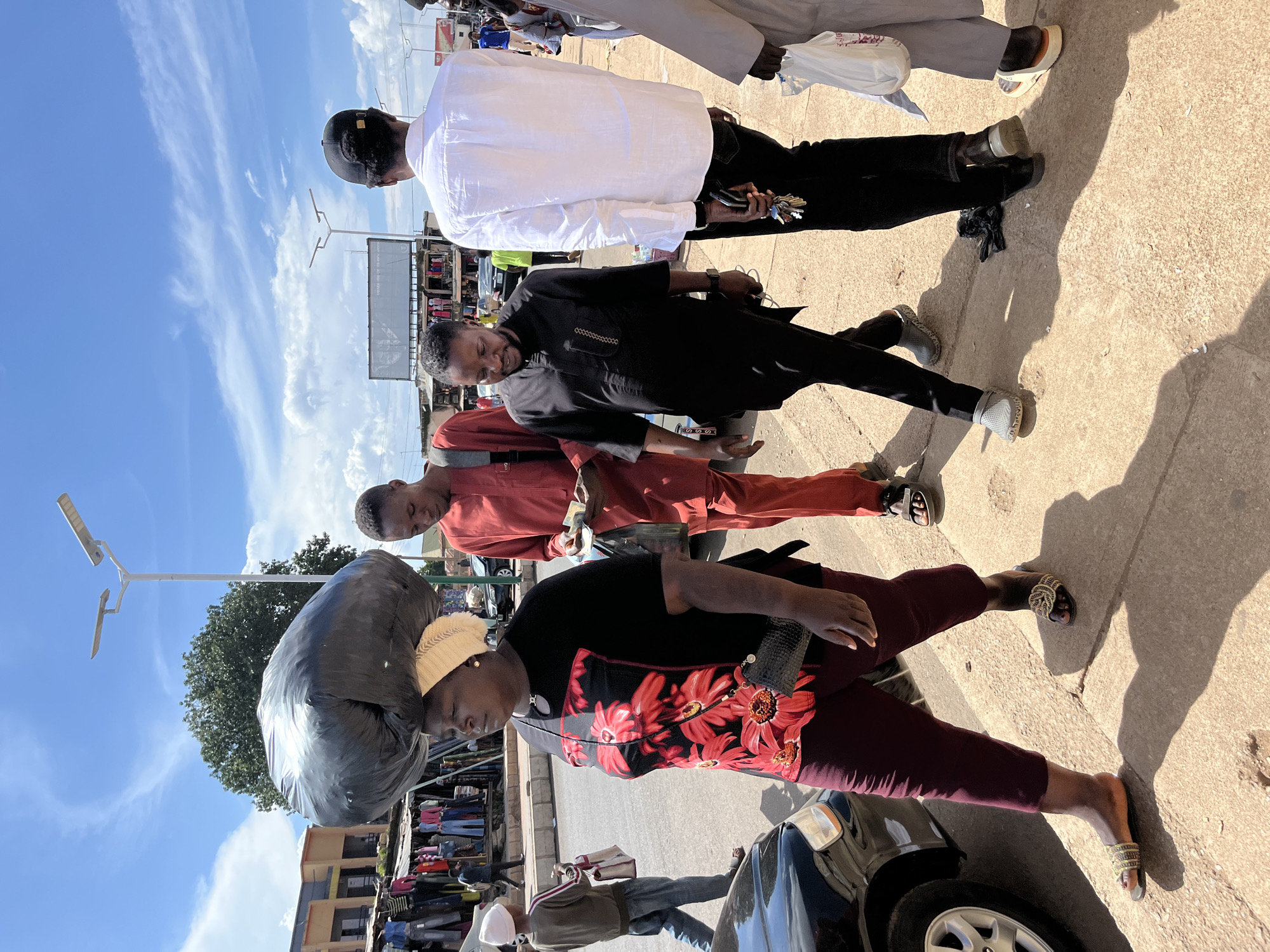 In the city of Jos, Nigeria, a woman walks on a sidewalk carrying a large black trash bag on her head. She is wearing a black t-shirt with red floral print and burgundy pants. To her right, a man in a red traditional outfit is walking, holding money in his hand. Further to the right, a man in a black traditional outfit is walking with his hands down by his sides. Behind them, another man in a white shirt and black pants is walking away from the camera. The street is lined with market stalls selling clothing. The sky is blue with scattered clouds. The scene appears to be a busy street in a commercial area.