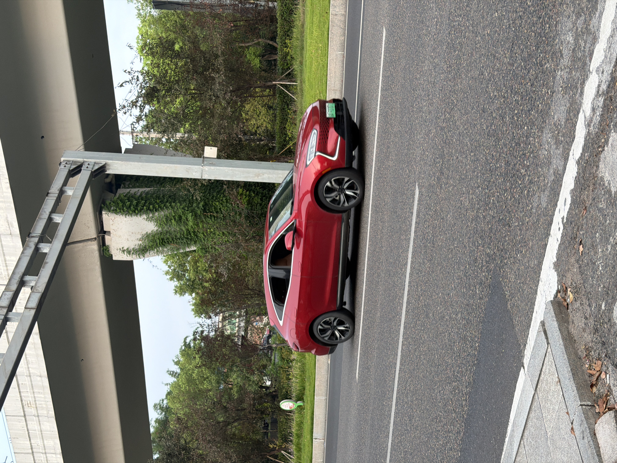 A red 2023 AITO M5 electric vehicle is driving on a road in Jiangjin, China. The car is a coupe SUV with a sporty design, featuring a sloping roofline and sharp body lines. It has a black grille with horizontal slats and distinctive LED headlights that wrap around the front corners. The car is equipped with multi-spoke alloy wheels and a green license plate.
The scene is set under an elevated highway structure, indicated by concrete pillars and a gray metal railing. Lush green trees and vegetation line the side of the road, with some trees showing darker foliage. The sky is overcast. The road surface is asphalt with white lane markings. A concrete sidewalk is visible along the edge of the road, with some fallen leaves scattered on it. The overall impression is a modern urban setting with elements of infrastructure and nature.
