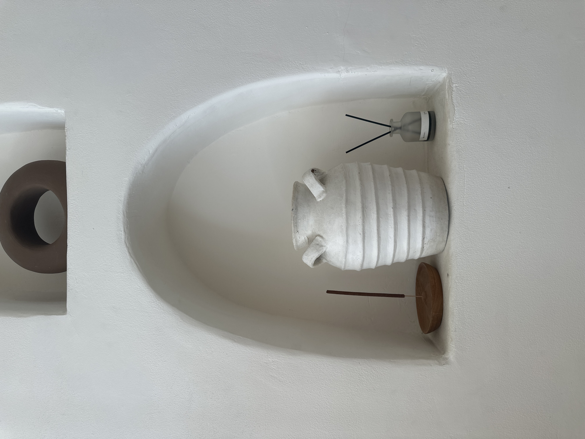 The image shows two recessed niches in a textured white wall, likely stucco. The location is North Kuta, Indonesia. The left niche contains a dark brown, circular, sculptural element. The larger, arched niche on the right houses a white, textured, amphora-style vase resting on a circular wooden base with a thin rod connecting the two. A reed diffuser in a clear glass bottle sits in the top right corner of the niche. The scene appears to be indoors, showcasing decorative elements within an architectural feature. There are no people visible in the frame.