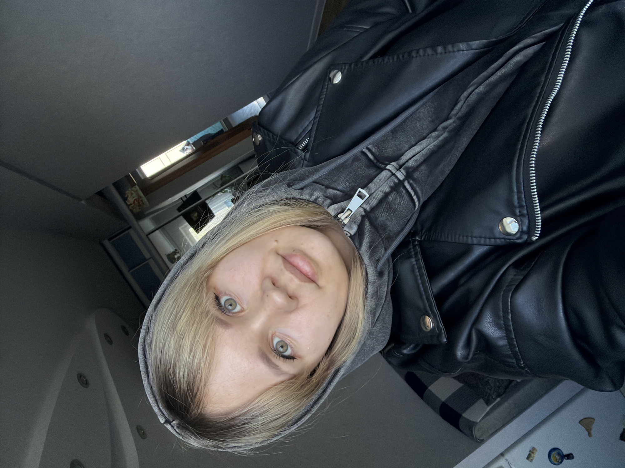 A dynamically angled, rotated close-up selfie features a young person with fair skin, blonde hair, and striking light green or hazel eyes. The subject looks directly into the camera with a neutral expression, their lips slightly parted. They wear a light grey hooded sweatshirt, with the hood pulled up, beneath a black faux leather jacket. The jacket features silver zippers and snap buttons, and its texture is visible with some creasing.
The background suggests an indoor setting with varied elements. To the upper left and center is a large, plain grey surface, possibly a ceiling or wall. In the middle-right, a window provides natural light, revealing a partial view of an outdoor scene with what appears to be blue sky and a distant window or object. A wooden frame outlines this window. In the lower left, a white fixture with several circular indentations or nozzles is visible, resembling part of a bathtub or jacuzzi. To the lower right, a white appliance, likely a refrigerator, is adorned with several small magnets, including one depicting a blue and white dish, a golden figure, and a pizza slice. A blue and white checkered fabric is draped over an unseen object next to the appliance. The lighting is a mix of natural light from the window and ambient indoor illumination, highlighting the subject's face. No discernible text is visible in the image.