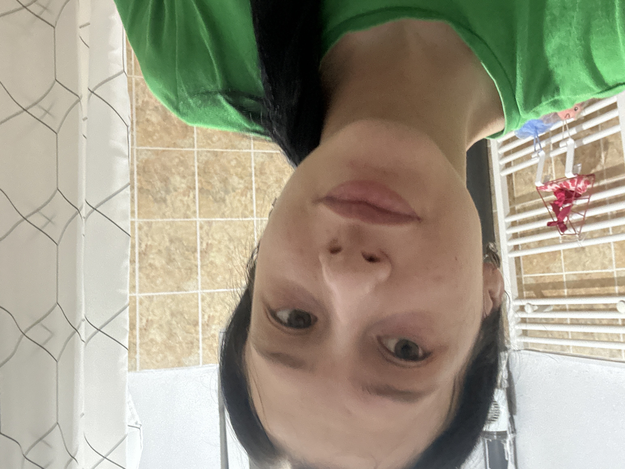 A young woman with dark hair, wearing a green t-shirt, is taking a selfie in what appears to be a bathroom. The background shows tiled walls, a heated towel rack with some items hanging on it, and a shower curtain. The lighting suggests an indoor setting. The woman's expression is neutral. The city of Perushtitsa, Bulgaria is not visible in the image.